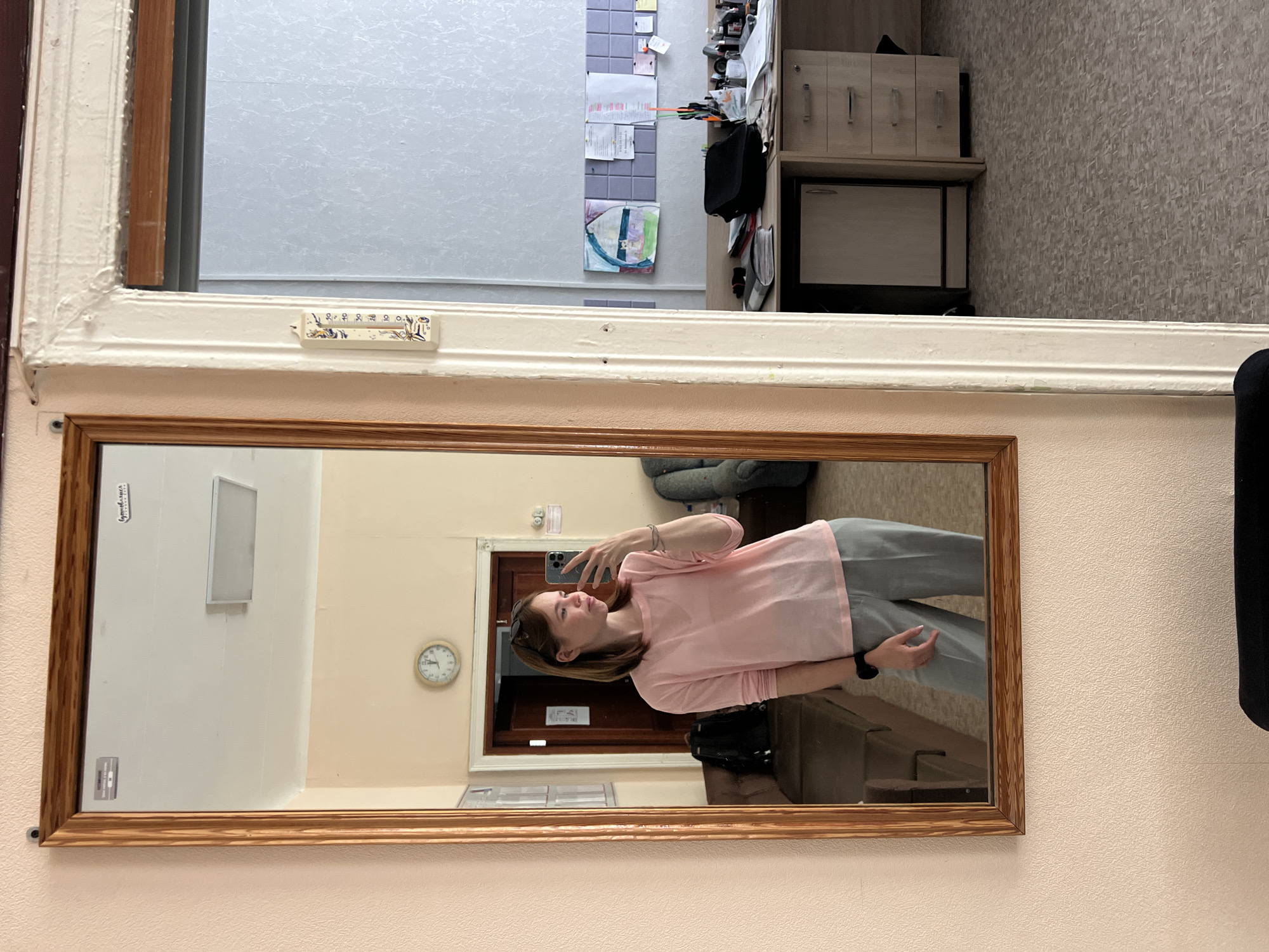 A mirror reflects a woman taking a selfie. She has shoulder-length brown hair, wears sunglasses atop her head, and is dressed in a pink top and gray pants. The mirror reflects an interior space with a clock on the wall, a closed wooden door, and furniture. The background out of the window shows an office space. The mirror is attached to a wall painted with a light pink color. There is a thermometer on the window frame above the mirror. The specific location within Chaikovsky, Russia cannot be definitively determined from the image.