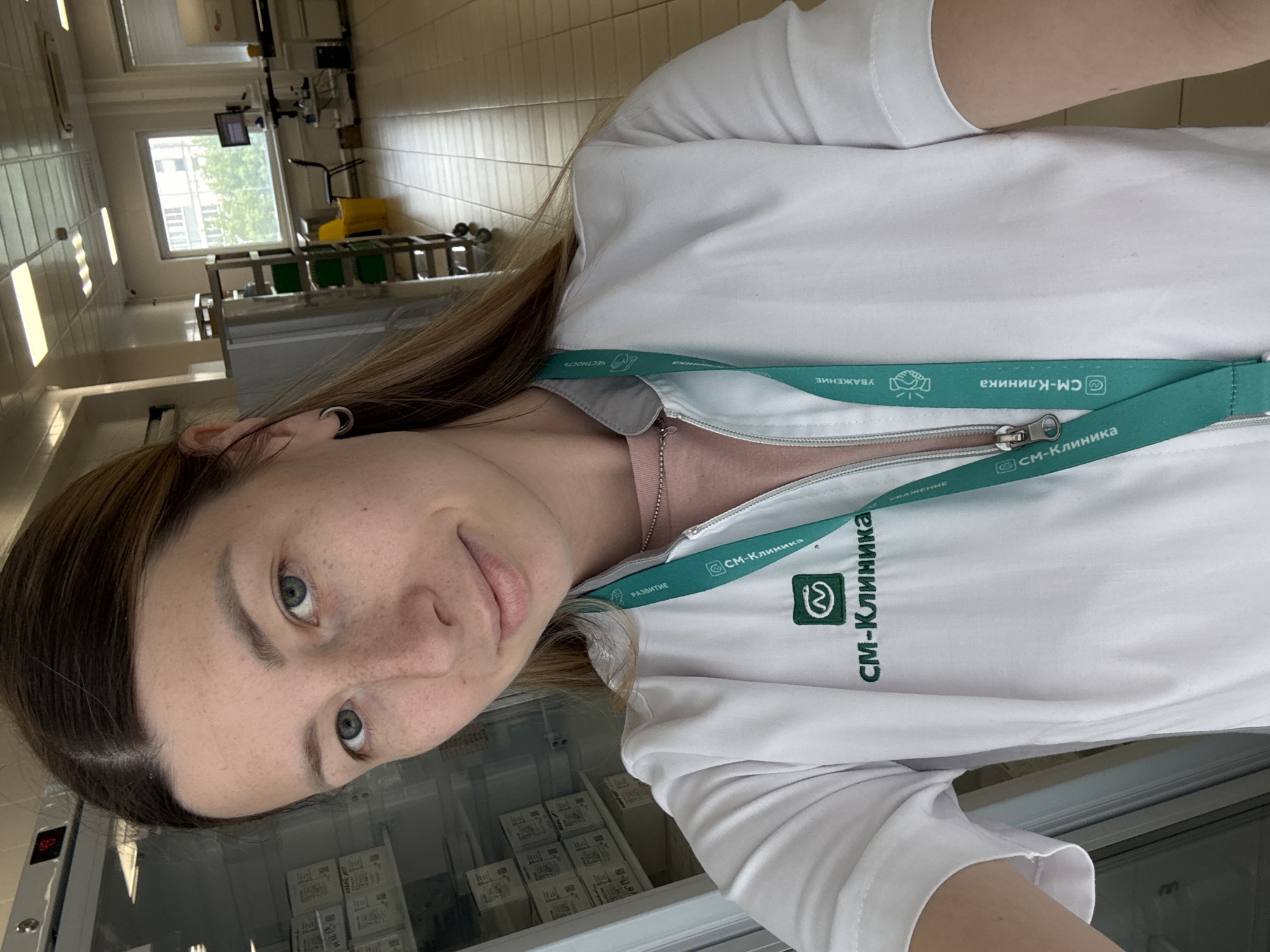 The image shows a young woman with light brown hair and blue eyes taking a selfie in a brightly lit indoor environment. She is wearing a white polo shirt with a green lanyard around her neck. The lanyard has text and logos visible, including "СМ-Клиника" (SM-Klinika), which appears to be a medical clinic chain. The background suggests a professional or medical setting, possibly a laboratory or clinic, with tiled walls and fluorescent lighting. Equipment and shelves are visible in the background, including what looks like medical devices and storage units. The woman has a faint smile on her face, and the overall mood is casual and perhaps work-related. The image is oriented vertically, as if taken with a phone held in portrait mode. The text "СМ-Клиника" is repeated multiple times on the lanyard and also appears as part of a logo on the shirt. Other visible text on the lanyard includes "УВАЖЕНИЕ" (Respect), "ЧЕСТНОСТЬ" (Honesty), and "РАЗВИТИЕ" (Development), which are likely company values. The setting appears to be indoors, and there are no indications of the time of day or weather conditions.