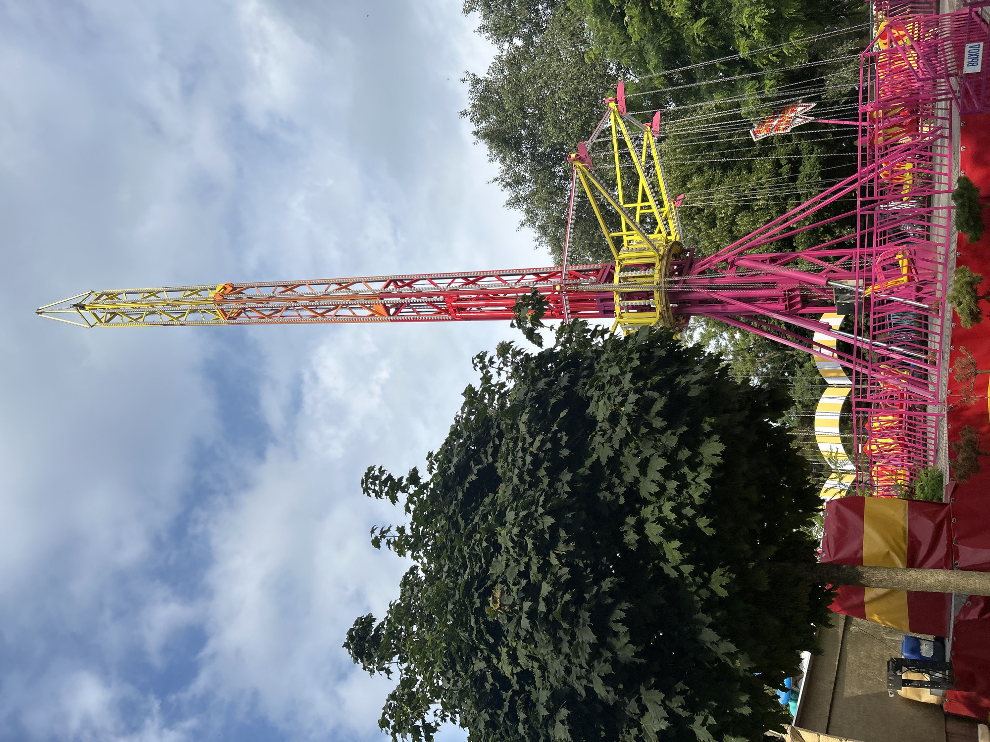 The image shows an amusement park ride with a tall, red and yellow tower structure. There are chains hanging from the tower, which appear to be part of a swing ride. The ride is surrounded by pink fencing and green trees. In the background, there is a blue sky with some clouds. The sign "ВЫХОД" (Exit) is visible on the pink fence. This amusement park is likely located in Gelendzhik, Russia.