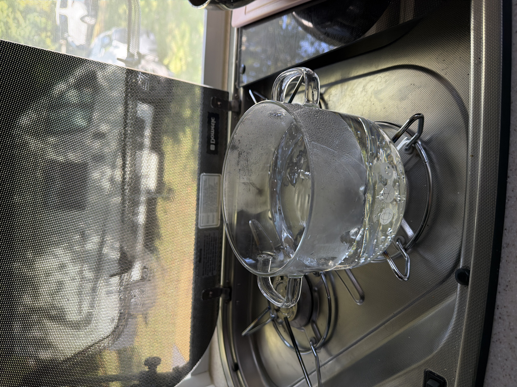 A glass mug filled with water is placed on a stovetop. The water appears to be heating up, with small bubbles visible on the surface and condensation on the glass. The stovetop is metallic and appears to be part of a mobile home or RV kitchen. In the background, a window is covered by a mesh screen, through which trees and vehicles are faintly visible. The scene suggests a domestic setting, possibly in Kazanlak, Bulgaria, given the context.
