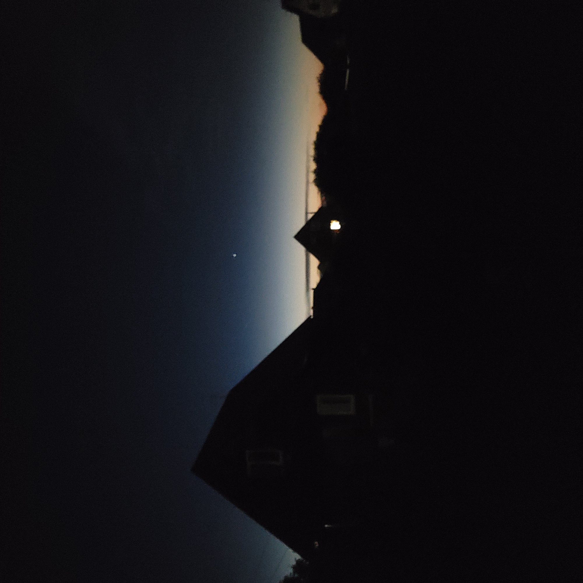 The image depicts a silhouette of houses against a dark twilight sky. The sky transitions from a deep blue at the top to lighter shades of blue and orange near the horizon. A single bright star or planet is visible in the upper portion of the sky. The houses are dark and indistinct, with some appearing to have lit windows. The overall scene conveys a sense of dusk or dawn in a rural or suburban setting.