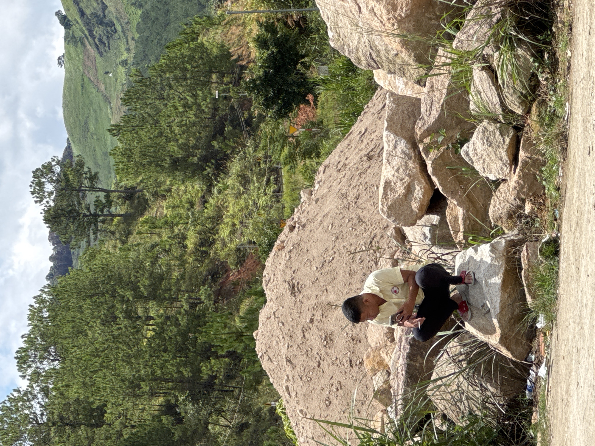 A man is squatting on a large rock, holding a mobile phone in his right hand. He is wearing a yellow short-sleeved collared shirt and black pants. The man is positioned in the foreground, with a large pile of sand behind him. In the background, there are steep, tree-covered hills. The environment appears to be rural or mountainous, with lush vegetation and a cloudy sky. There are some scattered rocks and grass around the man. The scene is consistent with the outskirts of Klong Klanh, Vietnam, a region known for its hilly terrain and agricultural or construction activities.