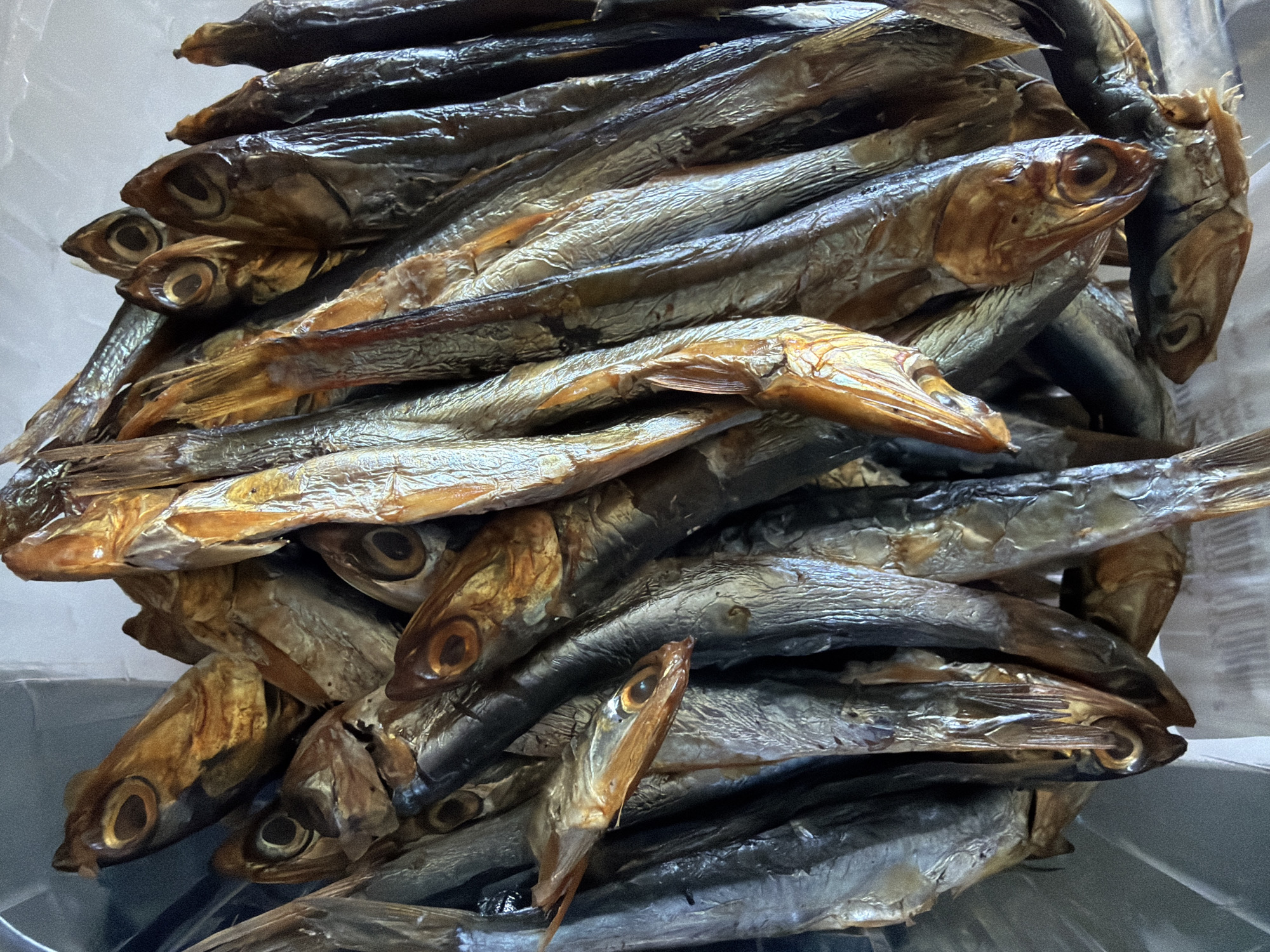 A close-up photograph shows a dense pile of small, whole, dried or smoked fish. The fish are arranged haphazardly, with many heads and bodies overlapping. The coloration of the fish varies from silvery-gray to brownish-gold, suggesting either different stages of preservation or different types of fish. The eyes of the fish are visible, some appearing dark and sunken. The fish appear to be displayed within a clear plastic container. The image lacks context to ascertain a specific location. Therefore, a location within Yerevan, Armenia, cannot be confirmed. There is no evidence of people, specific interactions, or environmental context within the provided image.