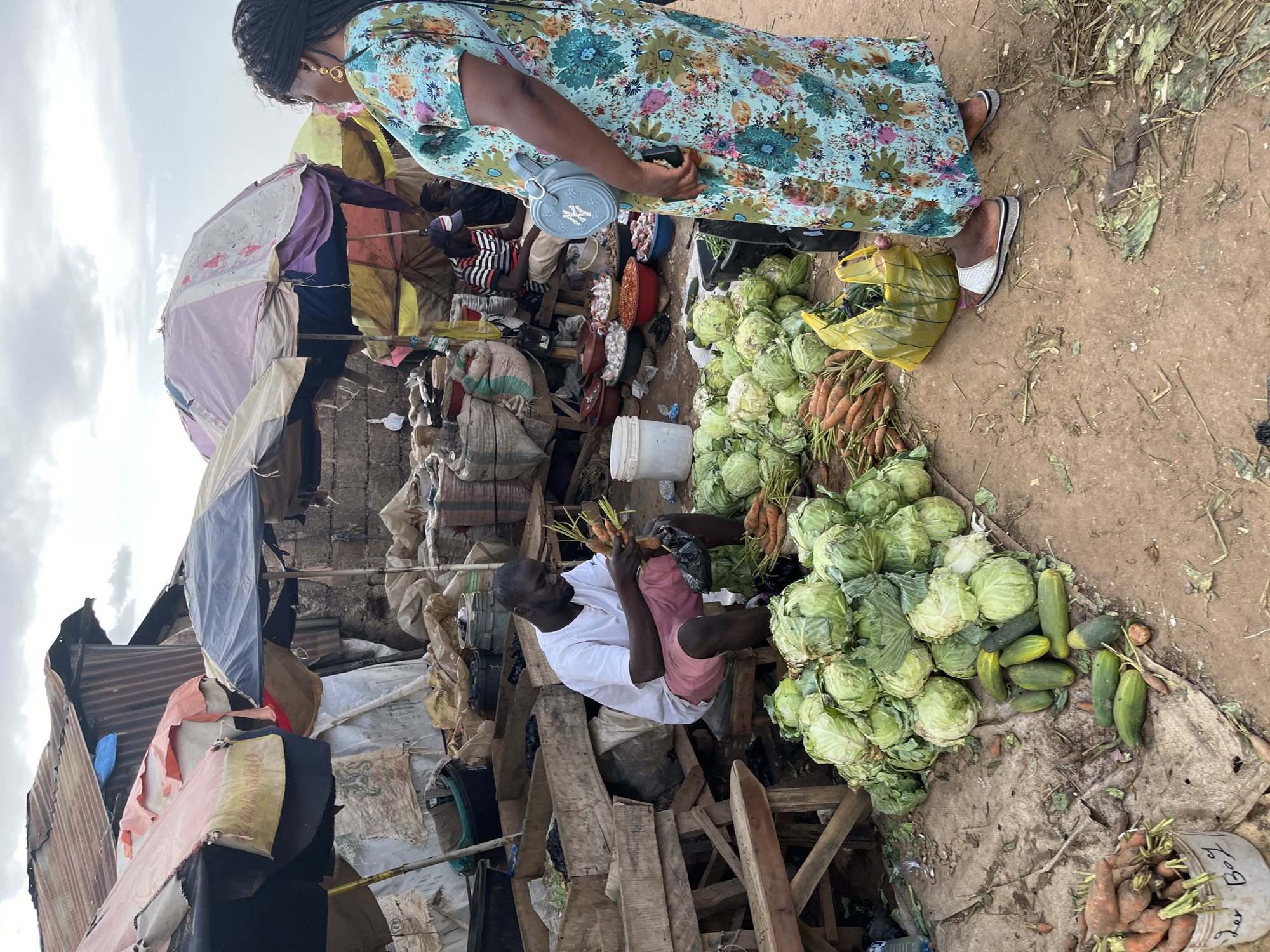 This image depicts an outdoor market scene in Rafin Par, Nigeria. In the foreground, a pile of green cabbages and cucumbers is arranged on the ground. To the left, a bucket filled with carrots is visible. A man in a white t-shirt and pink shorts sits on a stool, sorting through a bunch of carrots. In the background, several market stalls are set up with various goods. A woman in a floral dress stands to the right of the frame, facing away from the viewer. The environment appears to be a dusty, outdoor market with corrugated metal structures and umbrellas providing shade. The sky is overcast.