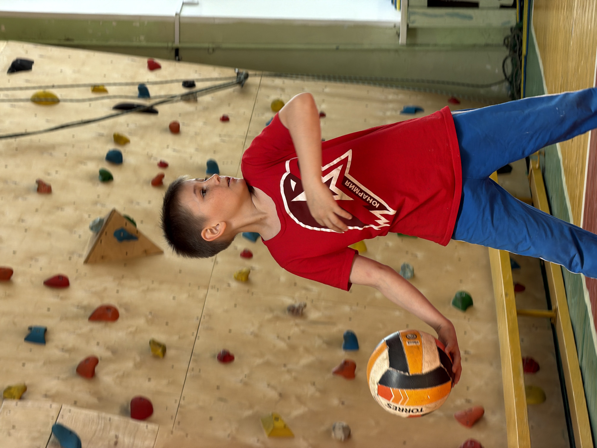 A young boy stands on a climbing wall in a building in Izhevsk, Russia. He is wearing a red t-shirt with white lettering and graphics on the front and blue sweatpants. He holds a multicolored soccer ball in his right hand. The climbing wall is light brown wood with various colored climbing holds attached. The floor is green and yellow. The photo is taken at a tilted angle.
