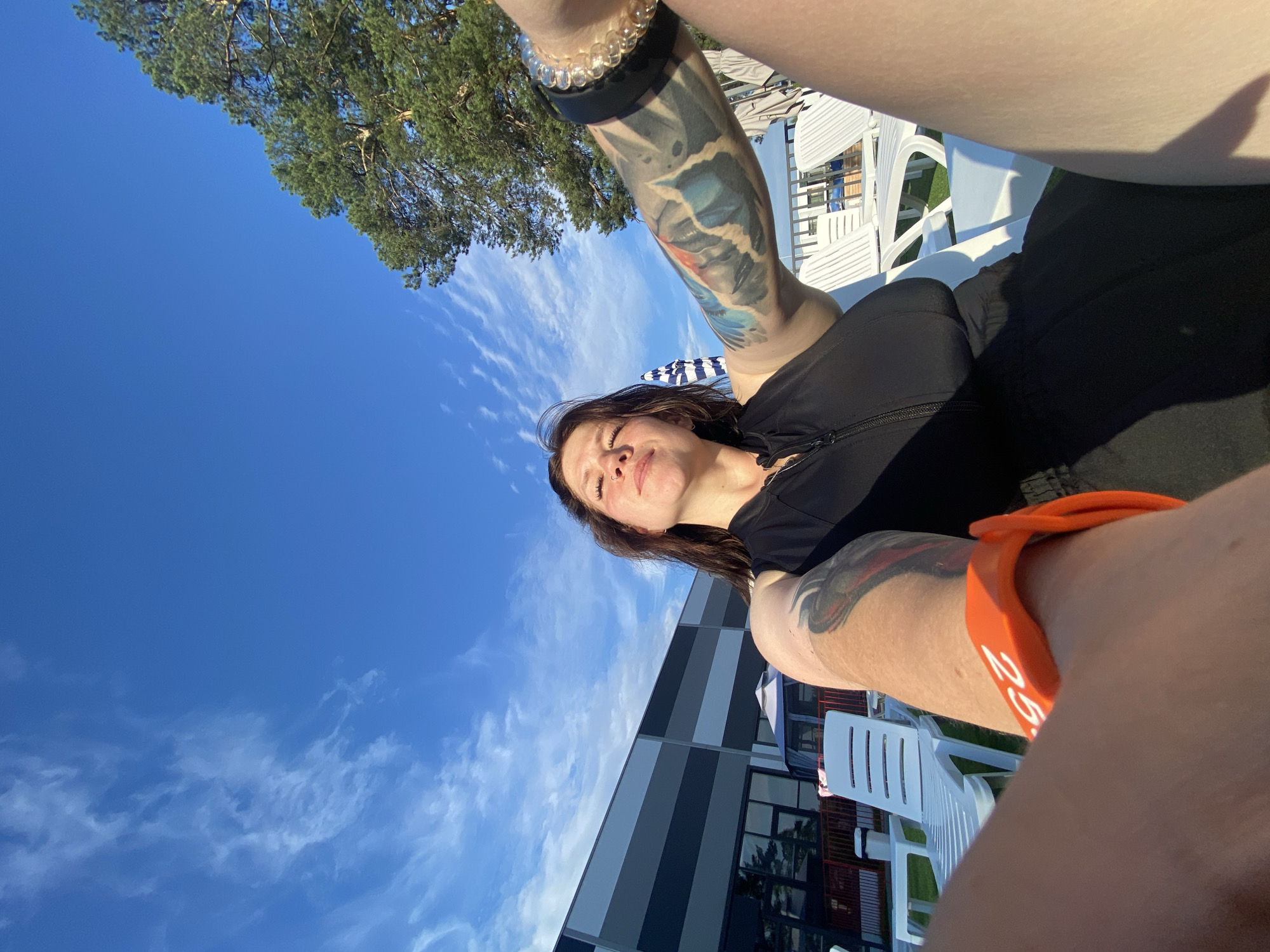 A woman is captured in a low-angle, selfie-style photograph outdoors on a sunny day, with a bright blue sky dotted with wispy white clouds above. She is sitting in a chair, her body angled towards the camera. She has long, dark hair, and her eyes are closed, with a slight smile on her face. She is wearing a black zippered top, and her left arm is visible, adorned with a large, colorful tattoo and a beaded bracelet. An orange wristband with the number "25" is also visible on her left wrist. In the background, there is a large building with horizontal grey and white stripes, likely a resort or hotel. Several white lounge chairs are arranged on a green lawn. A tall pine tree with lush green needles is visible on the right side of the frame. The scene appears to be set at a resort or recreational facility in Votkinsk, Russia, possibly near the Volga River, given the general landscape and architecture typical of such locations in the region.
