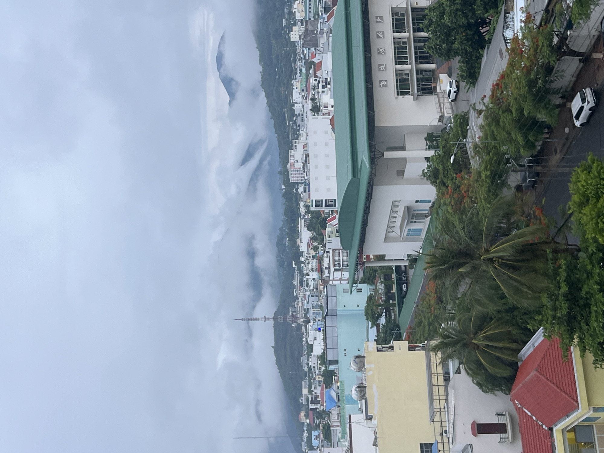 The image depicts a view of Bac Nha Trang Ward, Vietnam, from an elevated position, possibly a high-rise building. The sky is overcast with low-hanging clouds obscuring the mountain range in the background. The city is a mix of residential and commercial buildings, with various architectural styles and colors, including white, yellow, and light blue. There are green spaces interspersed among the buildings, with trees and palm trees visible. A street runs along the right side of the image with parked cars. There is a tall communication tower visible in the distance.