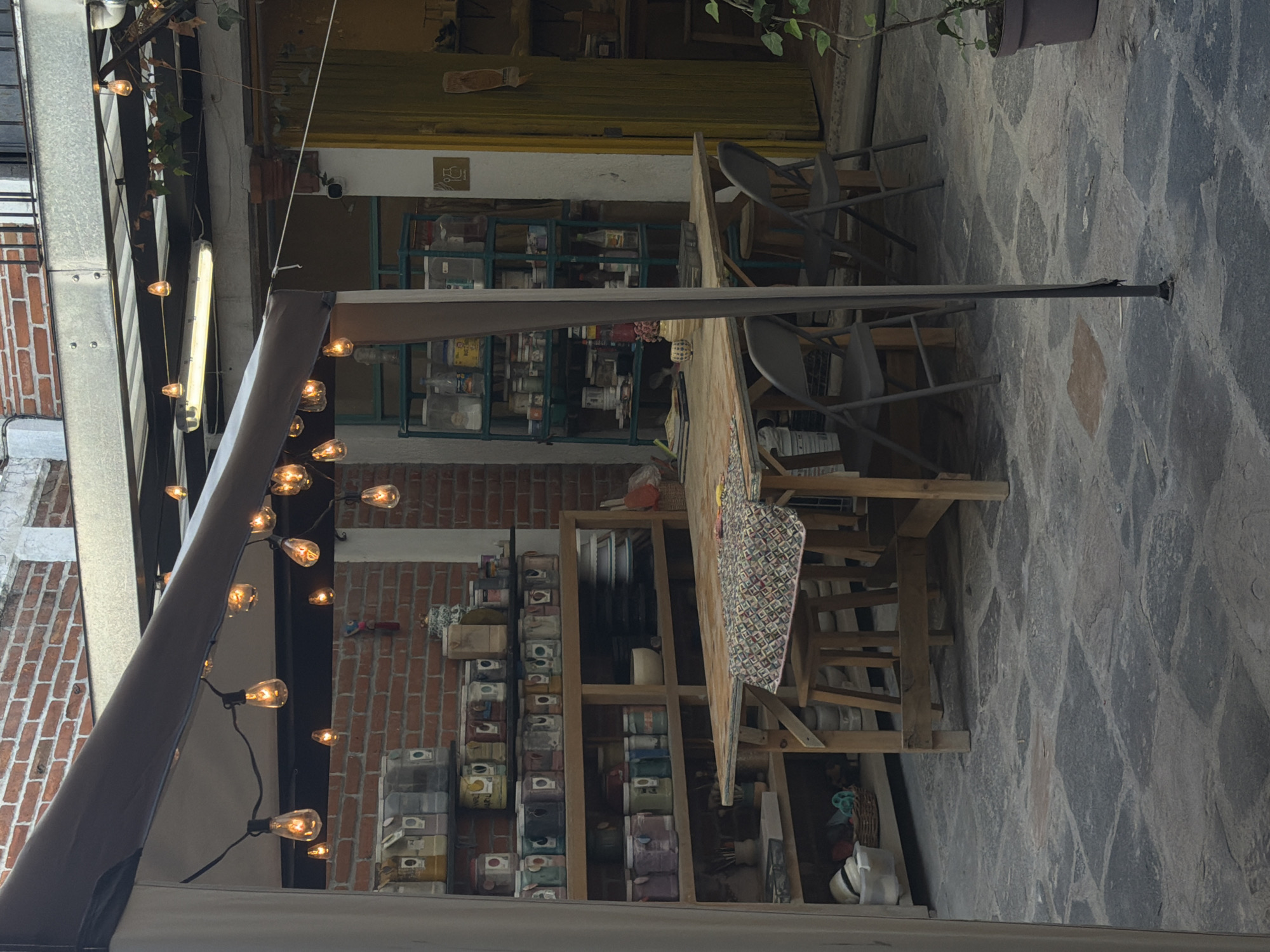 The image depicts a semi-enclosed outdoor or courtyard space, resembling a workshop or a rustic gathering area. At the center, a large wooden table is partially covered with a colorful patterned fabric, accompanied by several dark metal folding chairs. The area is illuminated by warm string lights with exposed bulbs, draped along an awning-like structure on the left, and a bright fluorescent tube light in the background.
Behind the table, various shelving units are visible. A tall wooden shelf to the right holds numerous jars and containers, while a metal shelf further back is filled with bottles and plastic containers. The background features brick walls, a segment of a white wall, and a prominent yellow door or shutter. A small white sign with a restroom pictogram (a person sitting on a toilet) is visible on the white wall section. A paintbrush hangs on the yellow surface above the metal shelves.
The ground is paved with irregular, dark-colored flagstones. A potted plant is seen in the upper right, and a white hat rests on the floor below the wooden shelves. The prominent artificial lighting suggests it is evening or night. No people are present, but the setup implies a functional workspace or a casual dining/gathering spot, with the containers hinting at storage for craft supplies, food, or other materials. The overall atmosphere is cozy and functional.
