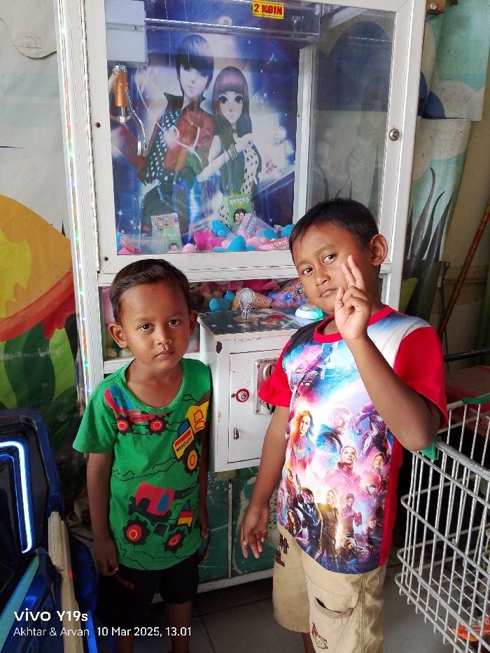 Two young boys are standing in front of a claw machine in a store in Kecamatan Haurgeulis, Indonesia. The claw machine has an image of two anime-style characters in the window. The boy on the left is wearing a green shirt with cartoon vehicles on it and has short, dark hair. He is looking down. The boy on the right is wearing a red and white shirt with cartoon characters on it and has short, dark hair. He is looking at the camera and has his right hand up near his face. The boys are both smiling. In the background, there is a shopping cart next to the claw machine. The store walls are covered in colorful designs. The image is taken with a cell phone camera. The time and date are displayed in the bottom left corner. The image is taken in the town of Kecamatan Haurgeulis in West Java, Indonesia, at coordinates -6.42 latitude and 107.955 longitude.