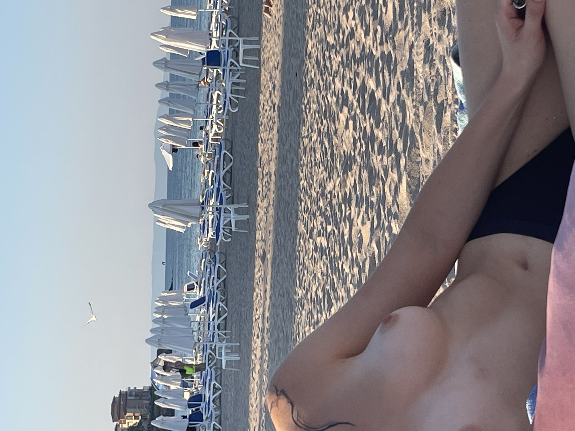 This media file captures a beach scene with a person in the foreground. The person, a woman, is lying down on the sand, with her torso and arm visible. She has a tattoo on her shoulder. Her face is not visible. In the background, there are rows of beach chairs and umbrellas, suggesting a populated beach. The sea is visible beyond the beach chairs, and a building can be seen in the distance. A bird is flying in the sky. The lighting indicates it is likely daytime, possibly late afternoon due to the long shadows cast by the beach chairs on the sand. The overall atmosphere appears relaxed and summery.