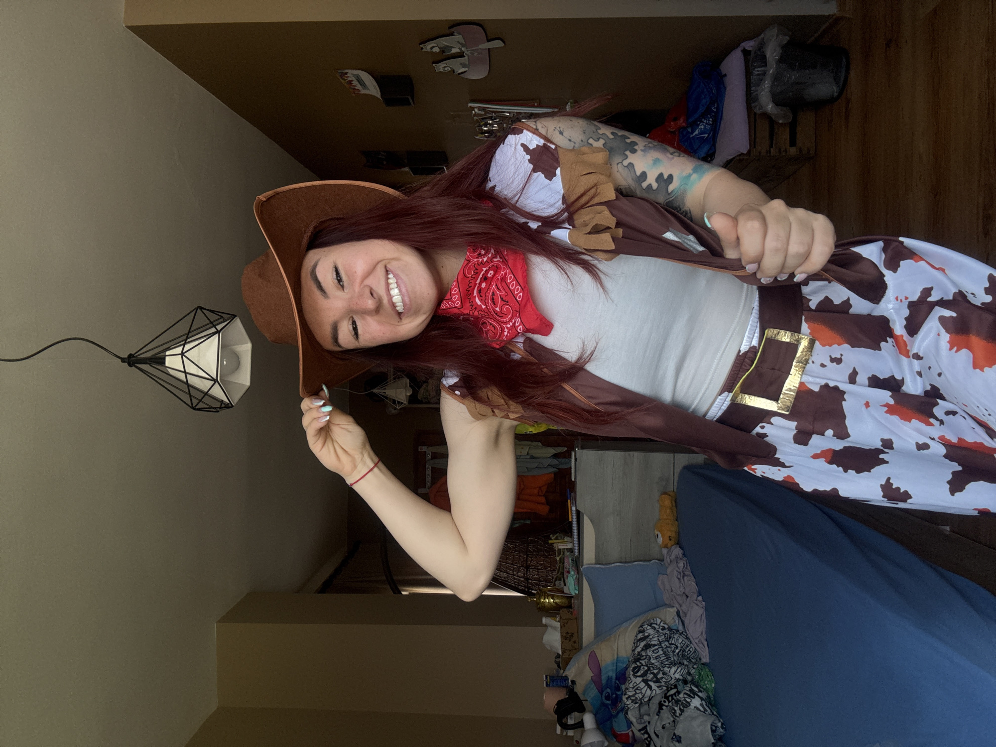 A young woman is posing in what appears to be an indoor setting. She is wearing a cowboy hat, a red bandana, a white top, and a cow-print dress with a brown vest. She is smiling and holding the brim of her hat. She has tattoos on her left arm. The room contains a bed with a blue comforter, a lamp with a geometric shade, and various objects in the background. There is no indication this photo was taken in the city of Ruse, Bulgaria.