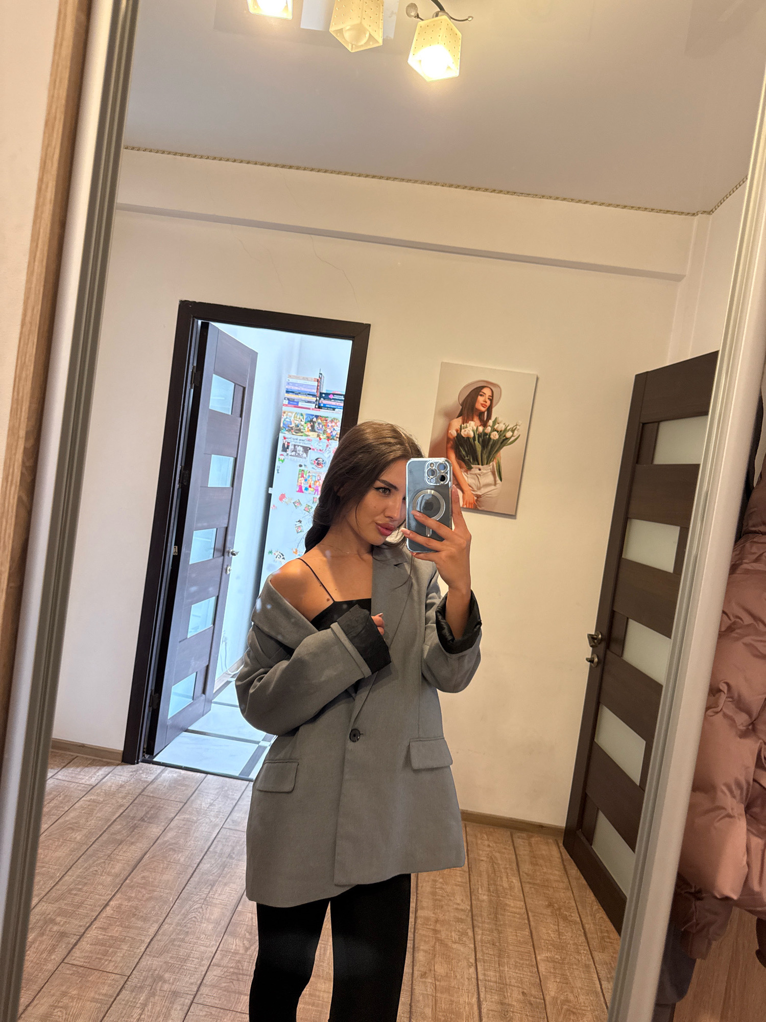 A young woman is taking a selfie in a mirror, standing in a room. She is wearing a grey blazer over a black top and black pants. Her left arm is over her shoulder, holding a smartphone. Behind her, a door with glass panels is slightly ajar, revealing a glimpse of what appears to be a hallway or another room. A framed picture of a woman holding tulips is hanging on the wall to her right, and a large mirror dominates the right side of the frame, reflecting part of the room, including a pink puffer jacket. The floor is made of wooden planks. The room appears to be an interior space, possibly a home or an apartment in Chișinău, Moldova.