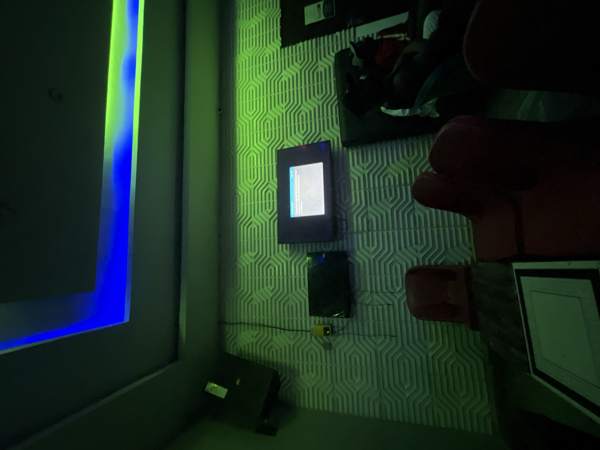 This image depicts an indoor entertainment space, likely a karaoke room or lounge, located in Jalingo, Nigeria. The room's illumination is primarily provided by artificial light, specifically green and blue LED strips recessed into a multi-tiered false ceiling. The green light casts a dominant hue across the room.
The main wall features white, geometric 3D panels arranged in an interlocking hexagonal or rectilinear pattern, strongly illuminated by the green light. A dark speaker cabinet is mounted high on the left side of this wall, angled downwards. Below the speaker, a small yellow control box with a green indicator light and visible wiring is affixed to the textured wall. Three flat-screen televisions are mounted on this wall: a smaller dark screen to the left, a larger central screen displaying a "no signal" message with white text on a dark background, and a partially visible dark screen to the far right. A small, dark, circular object, possibly a security camera, is visible near the ceiling above the central television. On the far right, a dark doorway or entrance is visible, with a white sign affixed to it or the adjacent wall.
In the foreground, a dark, low table with a central lighter-colored square insert is present. Two red/maroon upholstered chairs are positioned facing away from the camera, to the left of the table. On the right side of the room, two individuals are seated on dark, plush lounge seating. Their faces are obscured by shadow. One person, positioned further back, is wearing a red garment. The person closer to the foreground is wearing a cap and a lighter-colored shirt, with their legs extended.