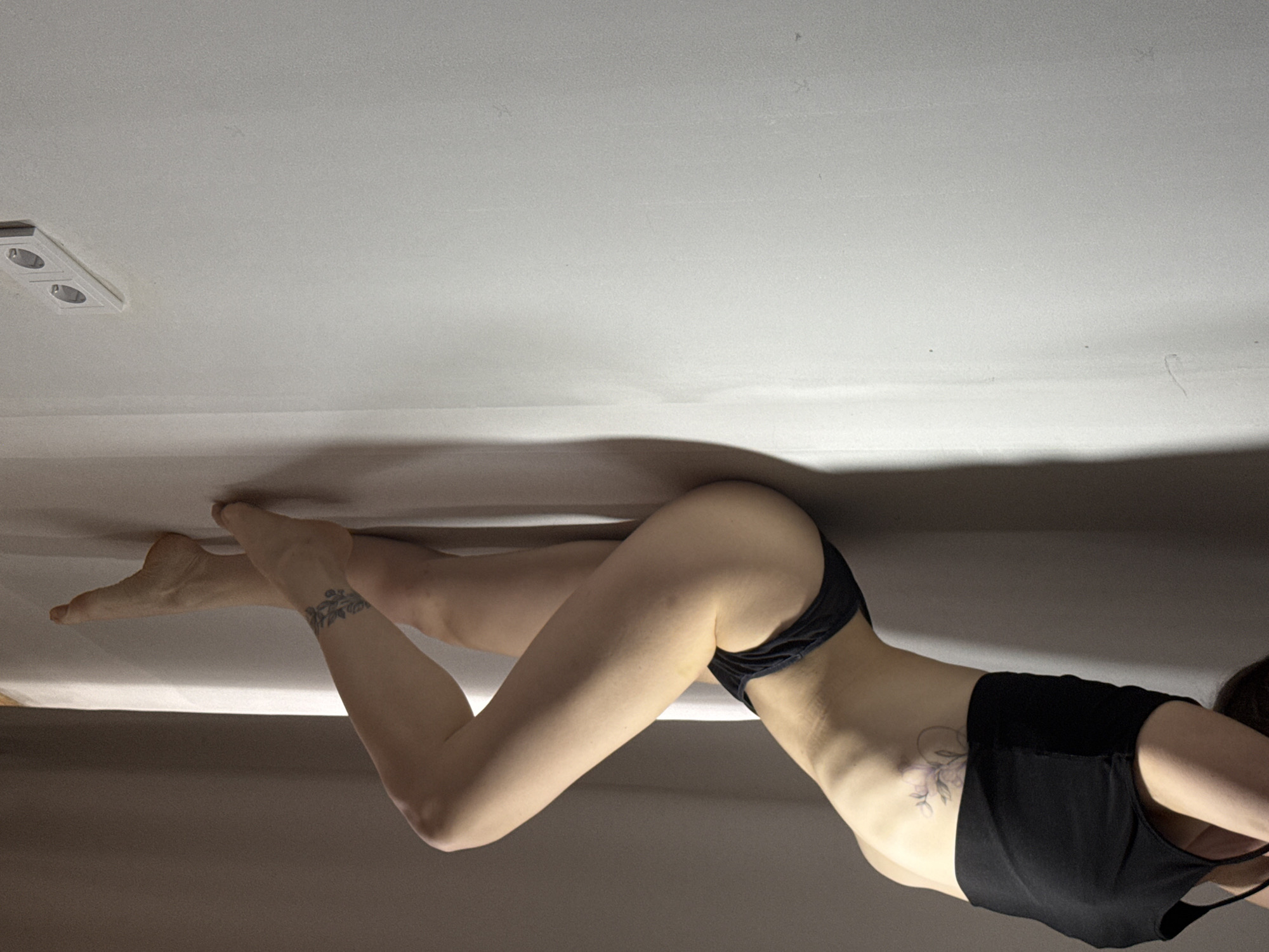 The image shows a woman lying on a bed. She is wearing black underwear and a black top. She has tattoos on her ankle and her side. The background is a plain white wall with a light fixture. There is no indication of the location being Chaikovsky, Russia.