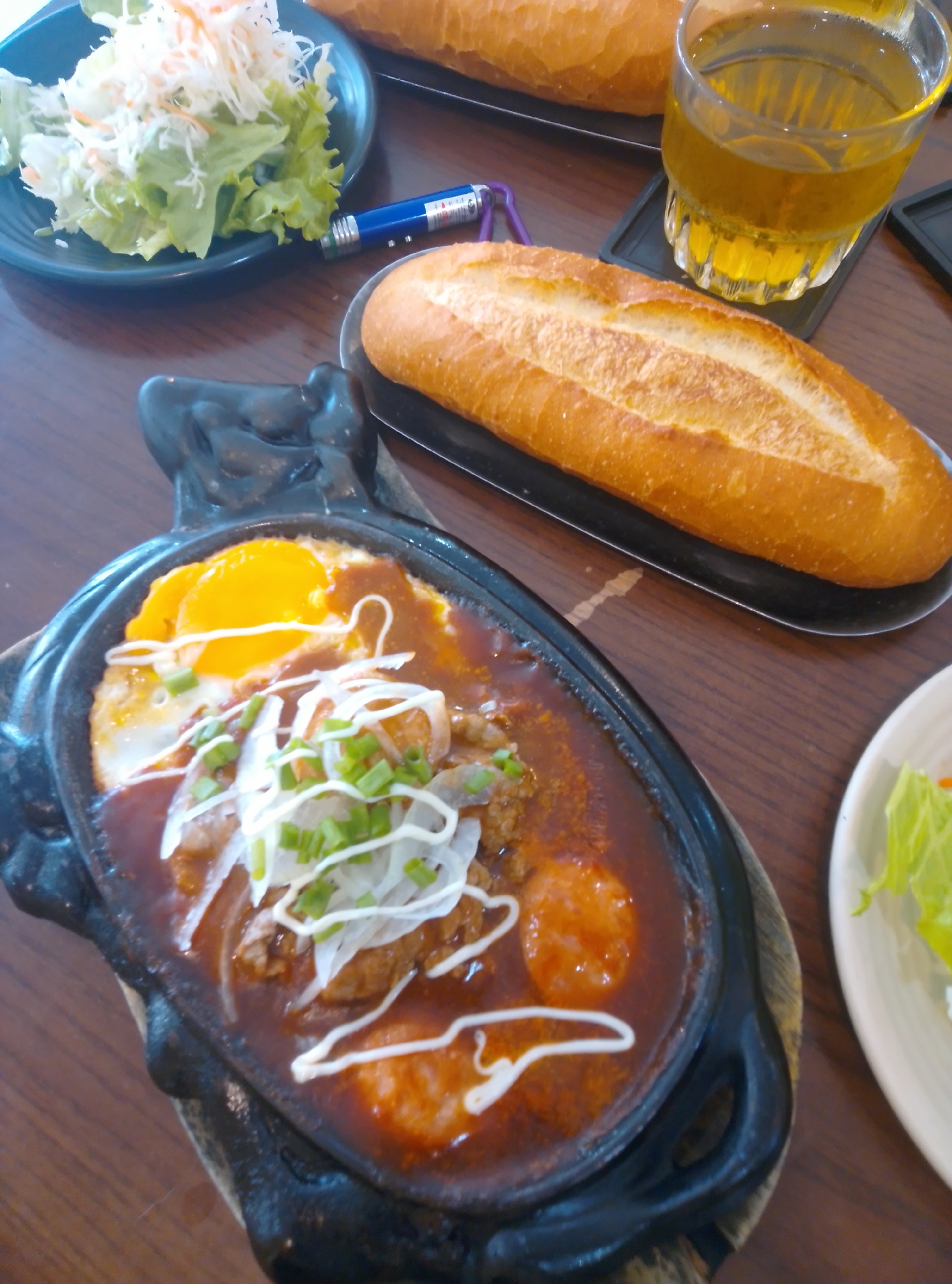 The image displays a meal set on a wooden table within an establishment in Bac Nha Trang Ward, Vietnam. The central focus is a black, oval-shaped sizzling plate, likely cast iron, resting on a wooden trivet. This plate contains a reddish-brown sauce, two fried eggs (one with an intact yellow yolk, the other with a partially broken yolk), pieces of cooked meat, and sliced white onions, all topped with chopped green onions and a drizzle of white sauce.
To the right of the sizzling plate, a golden-brown baguette is presented on a black, elongated dish. Another similar baguette is partially visible in the upper background, also on a black dish. In the upper left corner, a dark teal plate holds a fresh green salad composed of lettuce and shredded white and orange vegetables. Adjacent to this salad, a blue pen-shaped object, identified as a laser pointer due to its label and a visible purple lanyard, rests on the table.
In the upper right portion of the image, a clear glass filled with a light amber liquid and ice cubes, possibly iced tea or a similar beverage, sits on a black square coaster. A small, light-colored plate with additional green lettuce is partially visible on the far right edge of the frame. The wooden table surface exhibits a distinct grain pattern. No individuals are visible in the scene.
