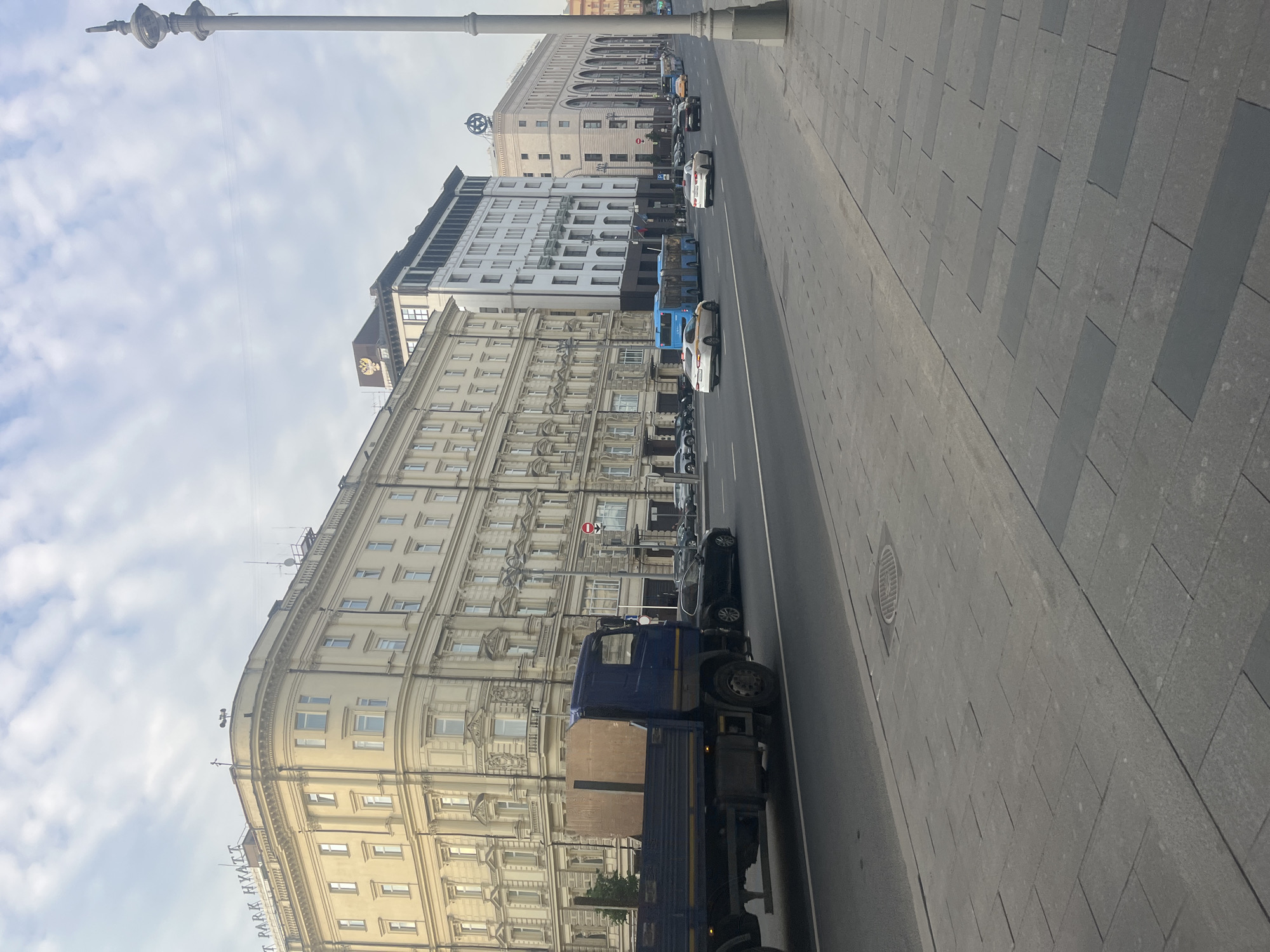A wide street scene in Moscow, Russia, features a row of large, ornate buildings on the left, with the "PARK HYATT" signage visible on the uppermost building. Several vehicles, including a blue truck with cargo, cars, and blue buses, are traveling on the multi-lane road. The sidewalk in the foreground is paved with grey rectangular stones. The sky is overcast with scattered clouds, and a streetlamp is visible on the right side of the image. The architectural style suggests a historic city center.