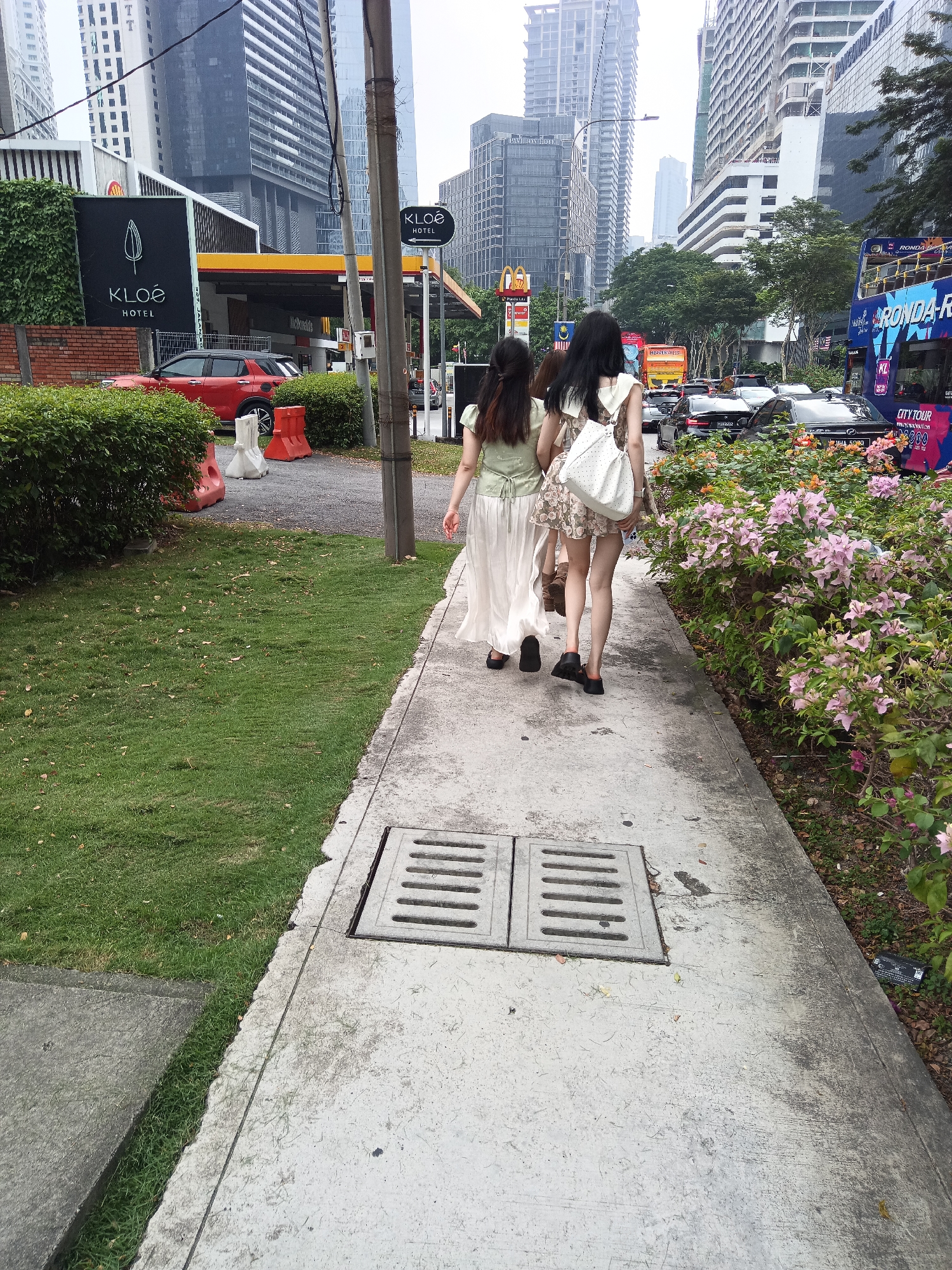 In Kuala Lumpur, Malaysia, two young women walk away from the camera on a concrete sidewalk adjacent to a grassy area. One woman wears a light green top and a long white skirt, and the other wears a floral print dress. Both are wearing dark shoes. Behind them, a street is visible with cars and a double-decker tour bus. Buildings and a gas station are also present in the background. The scene appears to be a public urban area.
