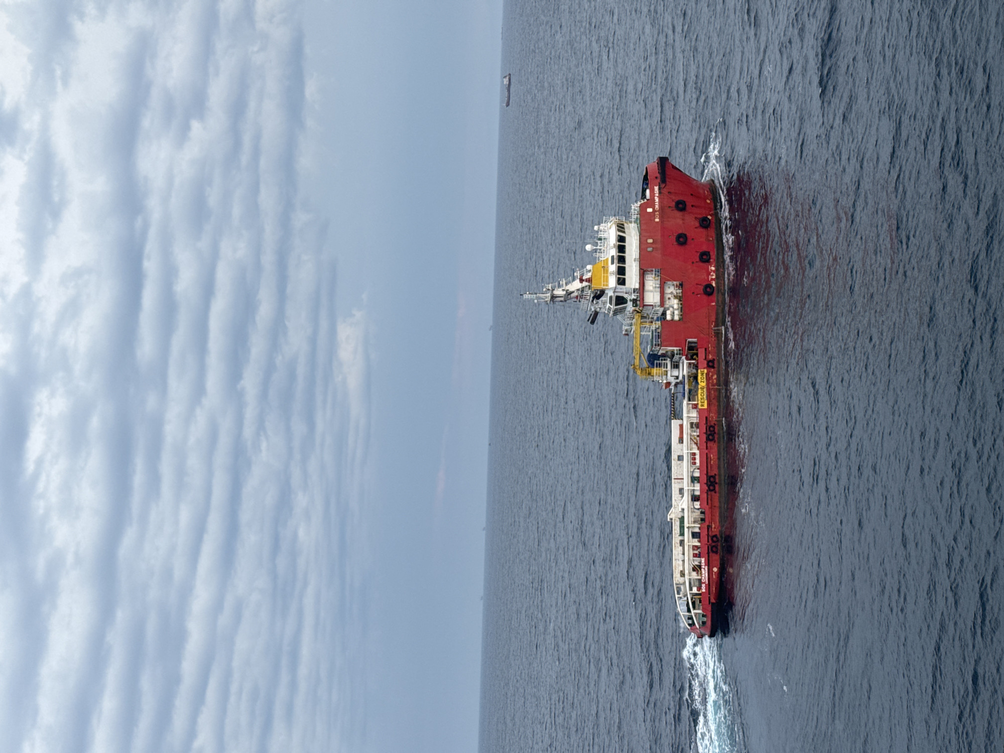 A red and white supply vessel, named "BOS CHAMPAGNE," is underway in a vast, choppy, dark blue ocean under a cloudy, overcast sky. The vessel is positioned in the center-right of the frame, leaving a white wake behind it. The ship has a tall white superstructure with yellow accents, and a red hull. The name "BOS CHAMPAGNE" is clearly visible on the side of the hull. There are several other ships visible on the horizon in the far distance, appearing as small, dark silhouettes against the lighter horizon. The overall scene suggests a maritime setting, likely at sea, with the vessel engaged in its operations. The location is not within a city, but rather in an open body of water.