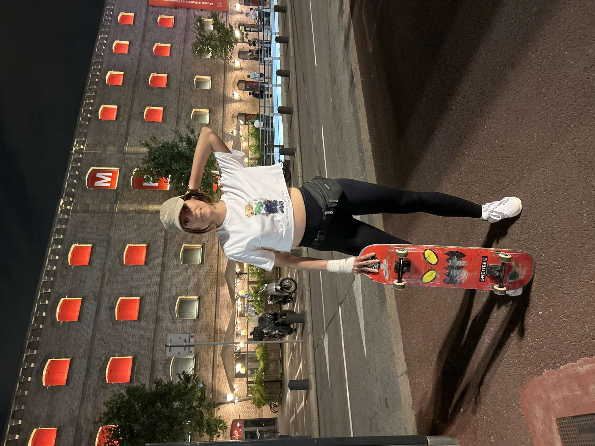 A person stands on a sidewalk at night in Barcelona, Spain, with a skateboard in hand. The person wears a white crop-top shirt, black leggings, a light-colored baseball cap, and white sneakers. Their left arm is wrapped in what appears to be a bandage or support. The skateboard is red with black and yellow designs. Behind the person, a large building with rows of illuminated red rectangular windows can be seen. The building has a sign with the letter "M" in a red square. The street is visible with parked motorcycles and an outdoor seating area with umbrellas. The lighting suggests it is night, with artificial lights illuminating the scene.