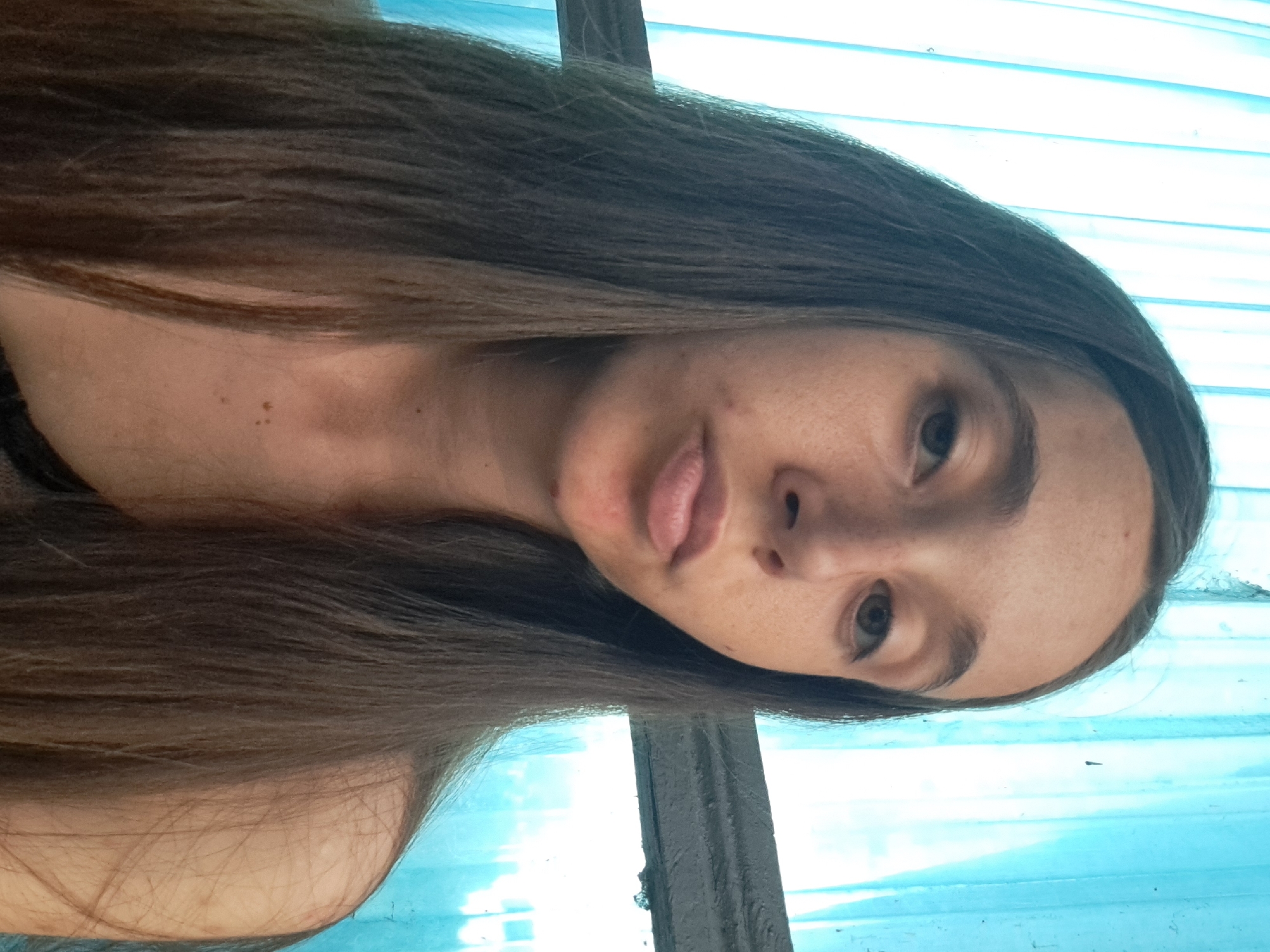 A young woman with long, dark brown hair is pictured from the chest up, looking directly at the camera. She has fair skin with freckles and a few blemishes visible. Her expression is neutral, with her lips slightly pursed. She is wearing a dark-colored garment that is partially visible at the bottom of the frame. The background consists of blue fabric that appears to be curtains or a similar material, with a horizontal gray bar or beam running across the middle of the image. The lighting suggests an indoor setting near a window, likely during the daytime. The overall scene is a close-up portrait.
