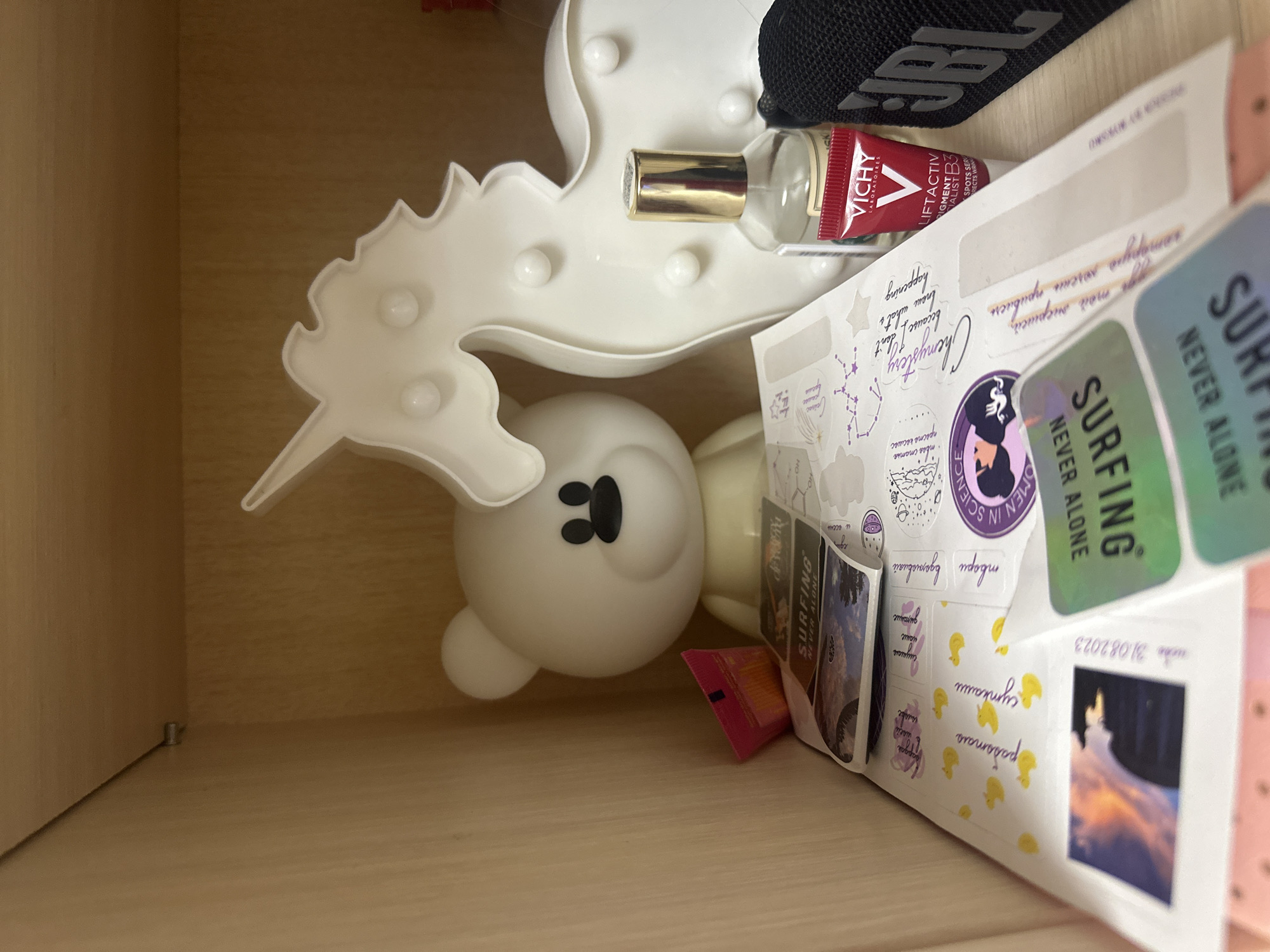 This media file captures a close-up, top-down view into what appears to be a light-colored wooden drawer or shelf in a personal space in Kazan, Russia. The contents are a mix of decorative items, personal care products, and a heavily stickered journal or scrapbook.
Dominating the left side is a white, seahorse-shaped light fixture with several small, round bulbs. Below it, a white, rounded figure resembling a bear or cartoon character, also appearing to be a light, is partially visible, lying on its side. To the top right, a black fabric item with the "JBL" logo is visible, likely a portable speaker or headphone case.
In the upper center, a small clear glass bottle with a golden cap stands near the seahorse light. Adjacent to it is a red and white tube of "VICHY LABORATOIRES LIFTACTIV PIGMENT SPECIALIST B3 SPOTS SERUM," indicating a skincare product.
The right half of the image is occupied by an open book or journal, filled with handwritten notes in both English and Cyrillic (likely Russian), alongside various stickers and small printed images. Prominent stickers include "SURFING NEVER ALONE" (repeated multiple times) and "WOMEN IN SCIENCE." Other stickers show a constellation pattern and small yellow ducklings. Handwritten English text reads "Chemystery because I don't know what's happening." Cyrillic handwritten words include "пачка" (pack), "сутками" (for days), "Слушаю" (I listen), "дихание" (breathing), "твори вдомовикі" (create at home), "твоя статья" (your article), "просто космос" (simply cosmos), "энершей" (energizer), and "привлечь" (to attract). A date, "31.08.2023," is clearly visible, along with a small printed image of an orange-hued cloud formation. A small pink sachet or packet is tucked beneath the papers.
The scene suggests a curated collection of personal items, possibly belonging to someone interested in beauty, music, and journaling. No specific activity is occurring; it is a static display of belongings.