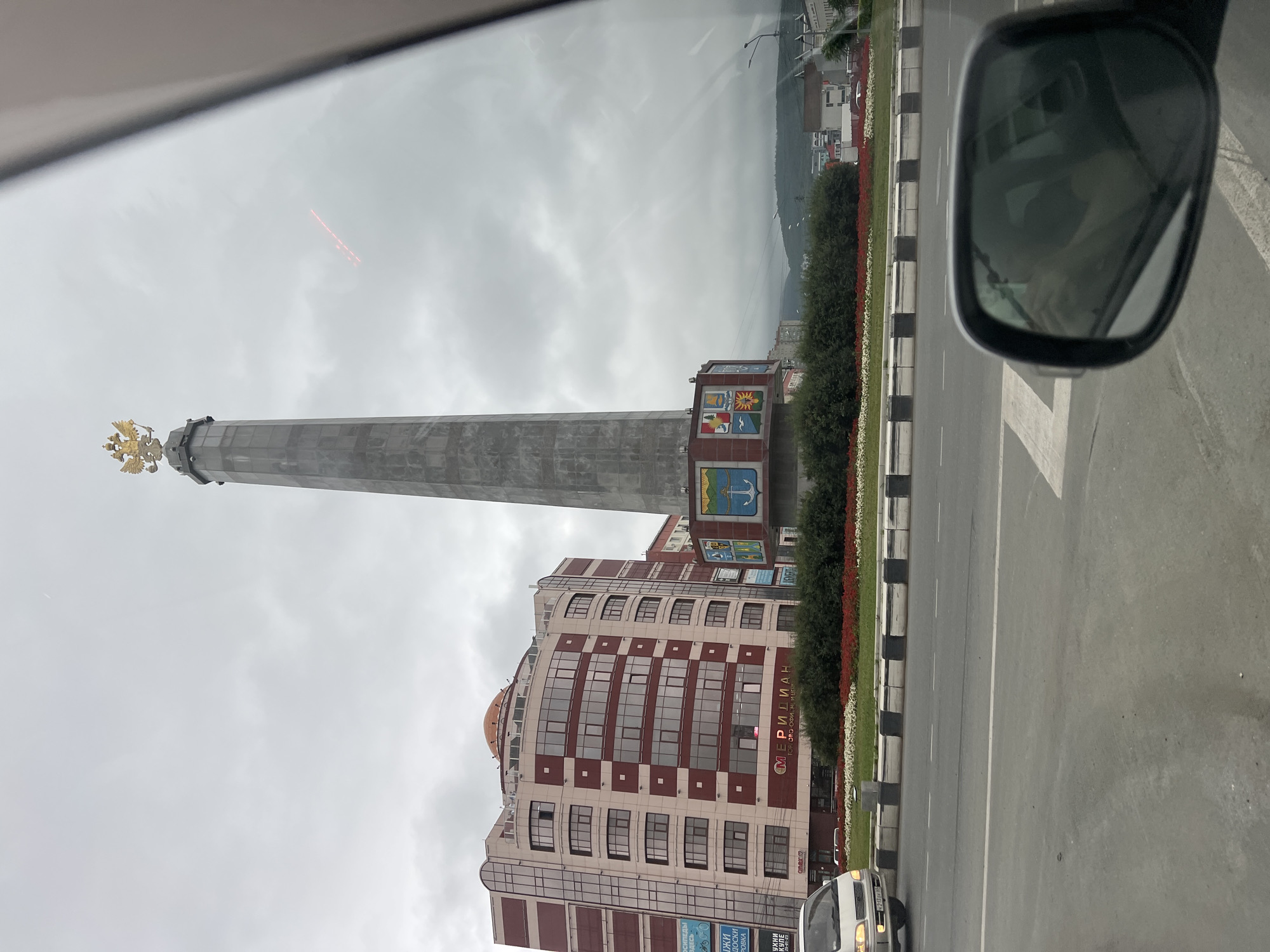A tall, grey obelisk topped with a golden double-headed eagle stands in the center of a roundabout in Yuzhno-Sakhalinsk, Russia. The obelisk is surrounded by a manicured garden with red and white flowers. To the left of the obelisk is a modern, multi-story building with a curved facade, featuring a pattern of red and beige panels and numerous windows. A sign on the building reads "Меридиан" (Meridian) and "ТОРГ. ОФИС. М. ЦЕНТР." (Trade. Office. M. Center). In the foreground, a grey road is visible, with a white van driving in the left lane. A car's side mirror reflects the interior of the car, showing a person's hand. The sky is overcast and cloudy, with hills visible in the background.