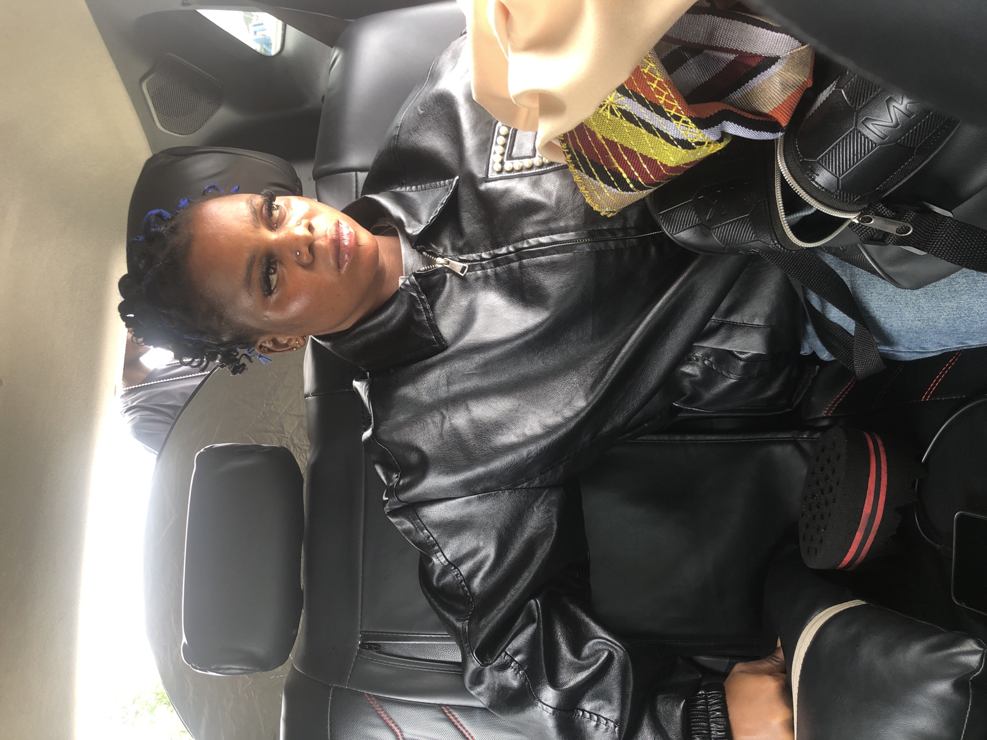In Ikoyi, Nigeria, a young Black woman sits in the backseat of a vehicle, wearing a black leather jacket and blue jeans. Her hair is styled in black braids with blue accents, and she has on makeup and jewelry. She is looking to her right, with her face slightly turned away from the camera. In her lap, she holds a small, patterned clutch and part of a beige garment. To her left, a black bag rests on the seat, and a black hair sponge is also visible. The interior of the car is dark, with black leather seats and visible seat stitching. A headrest is behind her, and the rear window is visible, with a bright outdoor light source illuminating the scene. Another person is partially visible in the background, wearing a grey shirt and a necklace.