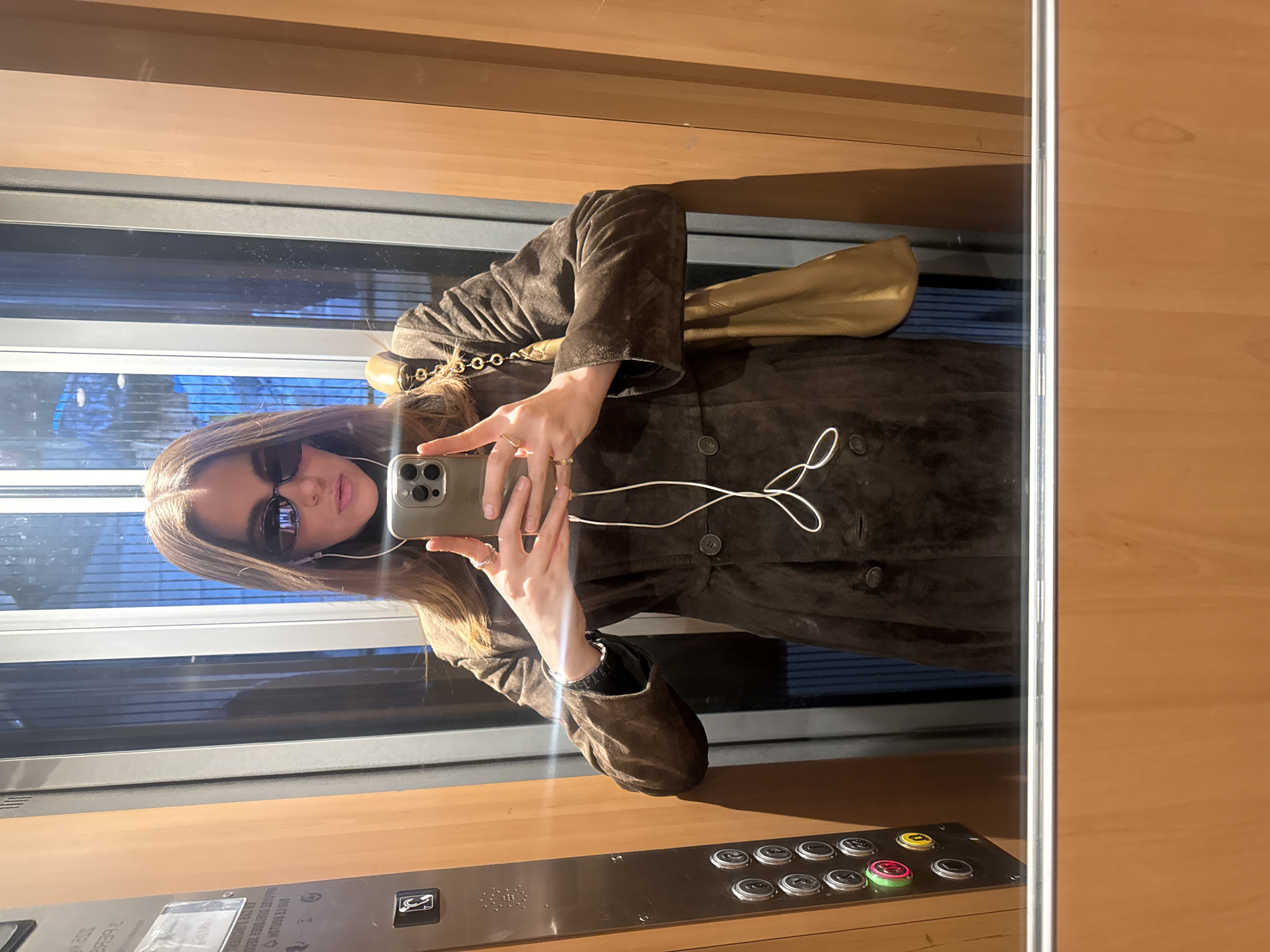 A person stands in an elevator, taking a selfie in the mirrored surface. The individual is wearing sunglasses and a brown suede-like jacket with buttons. A handbag with a gold chain is visible over the shoulder. The person is holding a smartphone, with a pair of wired earphones dangling. The elevator interior includes wood-paneled walls, a control panel with round buttons, and a window revealing the outside environment, which appears to be a cityscape with buildings visible through the glass. The location is Paris, France.