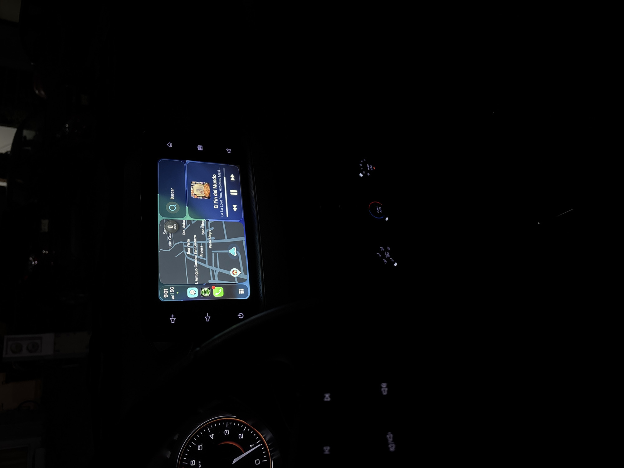 The image shows the interior of a vehicle, focusing on the infotainment system screen. The screen displays a map of San Francisco Ocotlán (Ocotlán), Mexico with navigation and a music player interface. The music player is playing "El Fin del Mundo" by La La Love You, Axolotes Mexicanos. Below the screen is the steering wheel, with only the speedometer visible. The clock on the screen indicates 9:01. The location on the map appears to be at or near "Antiguo Camino San Lorenzo" and "Riviera". The image is taken in a dark environment, with only the screen and some car dashboard lights illuminated.