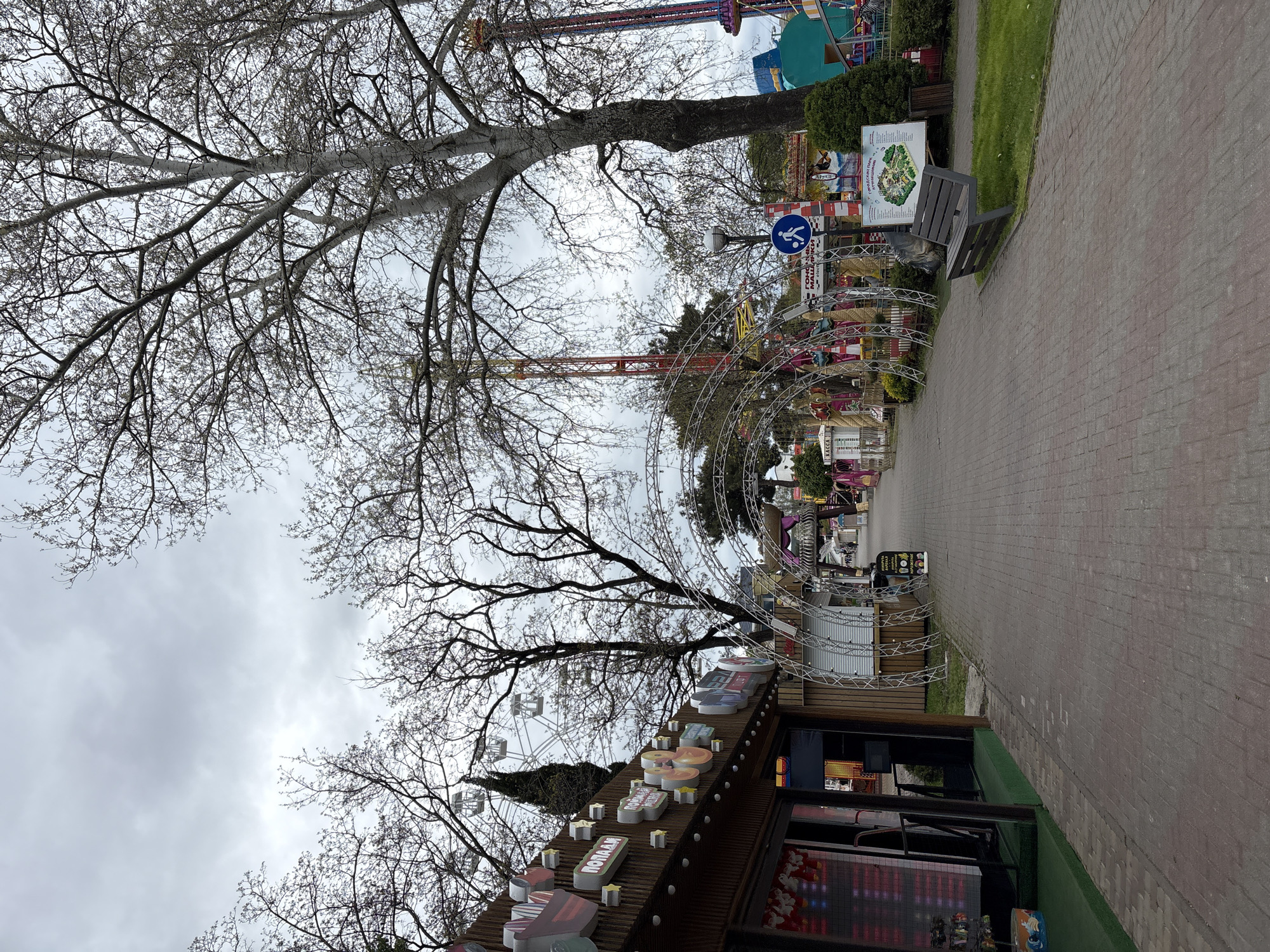 The image depicts a park setting in Gelendzhik, Russia. A paved brick pathway runs through the scene. Amusement park rides, including a Ferris wheel and a drop tower, are visible in the background. A metal archway with decorative elements lines the pathway. A tree with bare branches dominates the left side of the frame. To the right, a bench sits adjacent to the path. A kiosk or building with lit signage is on the lower left. A sign with information about the park is near the bench. The sky is overcast.