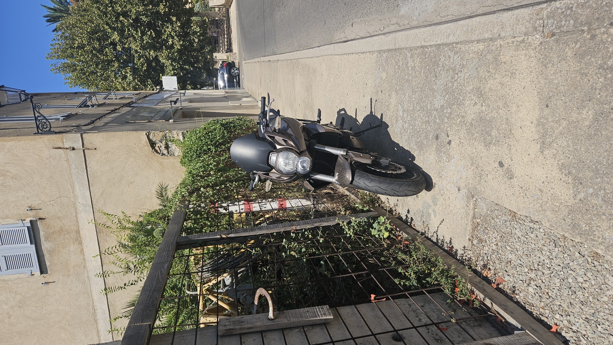 A brown motorcycle is parked on a sidewalk next to a street in L'Île-Rousse, France. The motorcycle has a dark windshield and is positioned facing forward, with its kickstand down. To the left of the motorcycle, there is a wooden and metal fence with overgrown greenery behind it. In the background, a light-colored building with blue shutters is visible, along with a large tree and a dark-colored car parked on the street. The sky is clear and blue.
