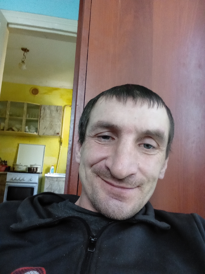 A male individual, appearing middle-aged with short dark hair and stubble, is depicted in a close-up selfie, smiling directly into the camera. He wears a dark, possibly black, zip-up hoodie over a grey garment. To the left and behind him, an open doorway reveals an adjacent room, identified as a kitchen in a residence located in Yarun, Ukraine. This room features vibrant yellow walls, a white ceiling with a light blue upper border, and a hanging light fixture with an exposed bulb. Visible kitchen elements include two wall-mounted cabinets with translucent doors, a white stove/oven with two pots on its surface, and a sink area with a faucet. A circular vent is present on the yellow wall. The subject occupies the majority of the foreground and right side of the frame, partially obscuring the wooden door or wall panel behind him on the right.
