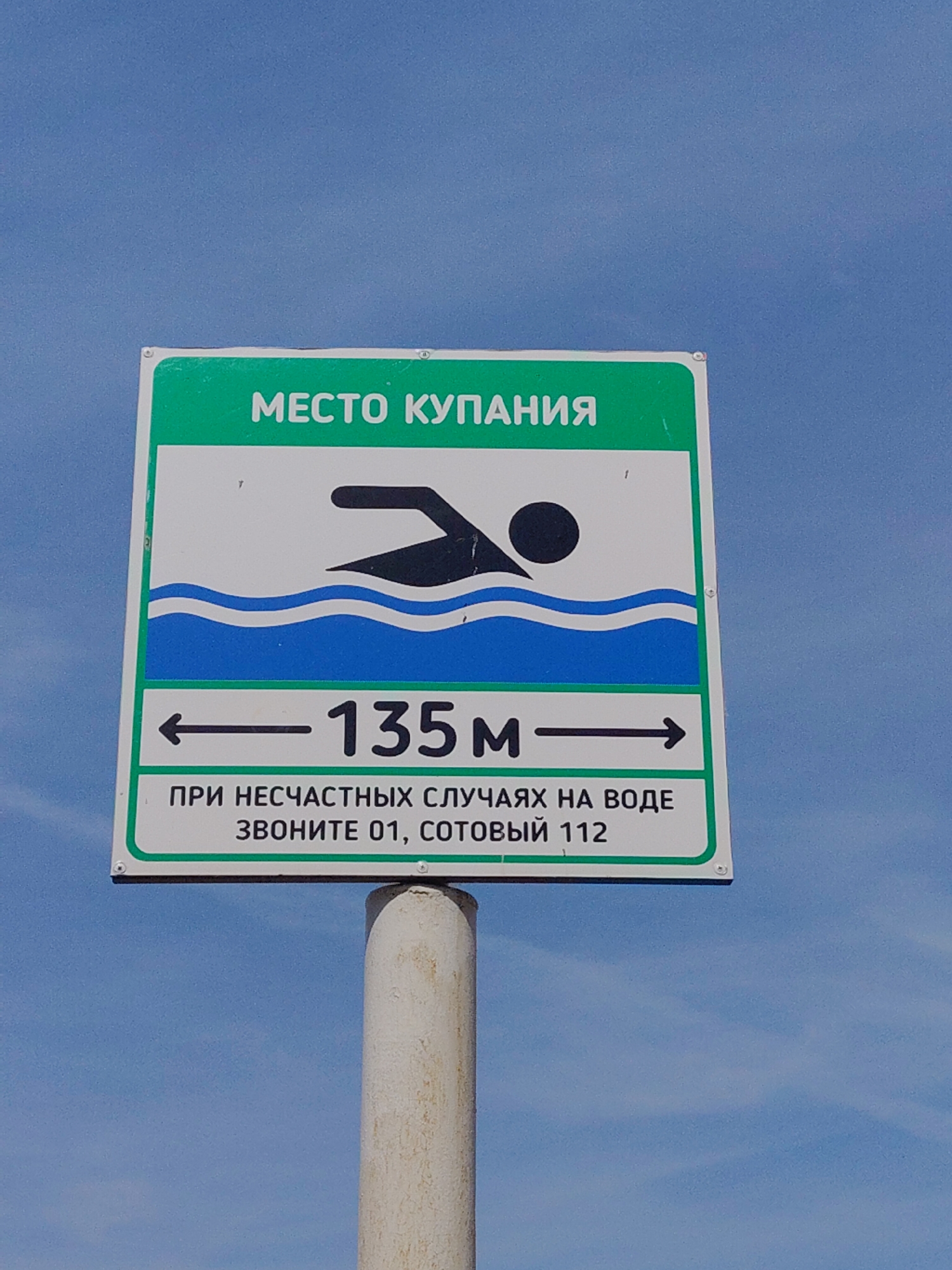 The image shows a square sign with a green border against a clear blue sky. The sign depicts a black silhouette of a swimmer in blue water waves. Above the swimmer, the sign reads "МЕСТО КУПАНИЯ" in green letters, which translates to "Swimming Place" in English. Below the swimmer, there is a horizontal arrow pointing both left and right with the text "135M" in between. At the bottom of the sign, there is text that reads "ПРИ НЕСЧАСТНЫХ СЛУЧАЯХ НА ВОДЕ ЗВОНИТЕ 01, СОТОВЫЙ 112", which translates to "In case of accidents on the water call 01, mobile 112". The sign is mounted on a white, cylindrical post. The location is presumed to be a swimming area, but the city and specific location are unknown.