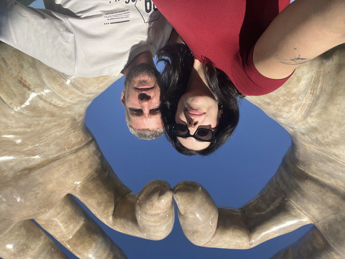 A selfie photograph captures a female and a male subject positioned in front of the "Hands of Love" sculpture at Hon Chong Promontory in Nha Trang, Vietnam. The prominent sculpture in the background features two oversized, beige-toned hands with a glossy finish, forming a heart shape against a clear blue sky.
The female subject, on the left, has dark hair and wears black sunglasses and a red short-sleeved top. Her left forearm, extended towards the camera, displays a small, dark tattoo. She is looking directly at the camera.
The male subject, on the right, has short, light grey hair and a beard. He wears a light grey long-sleeved crew-neck shirt featuring a black graphic print on the lower front. He is also looking directly at the camera. Both individuals are partially illuminated by direct sunlight.
