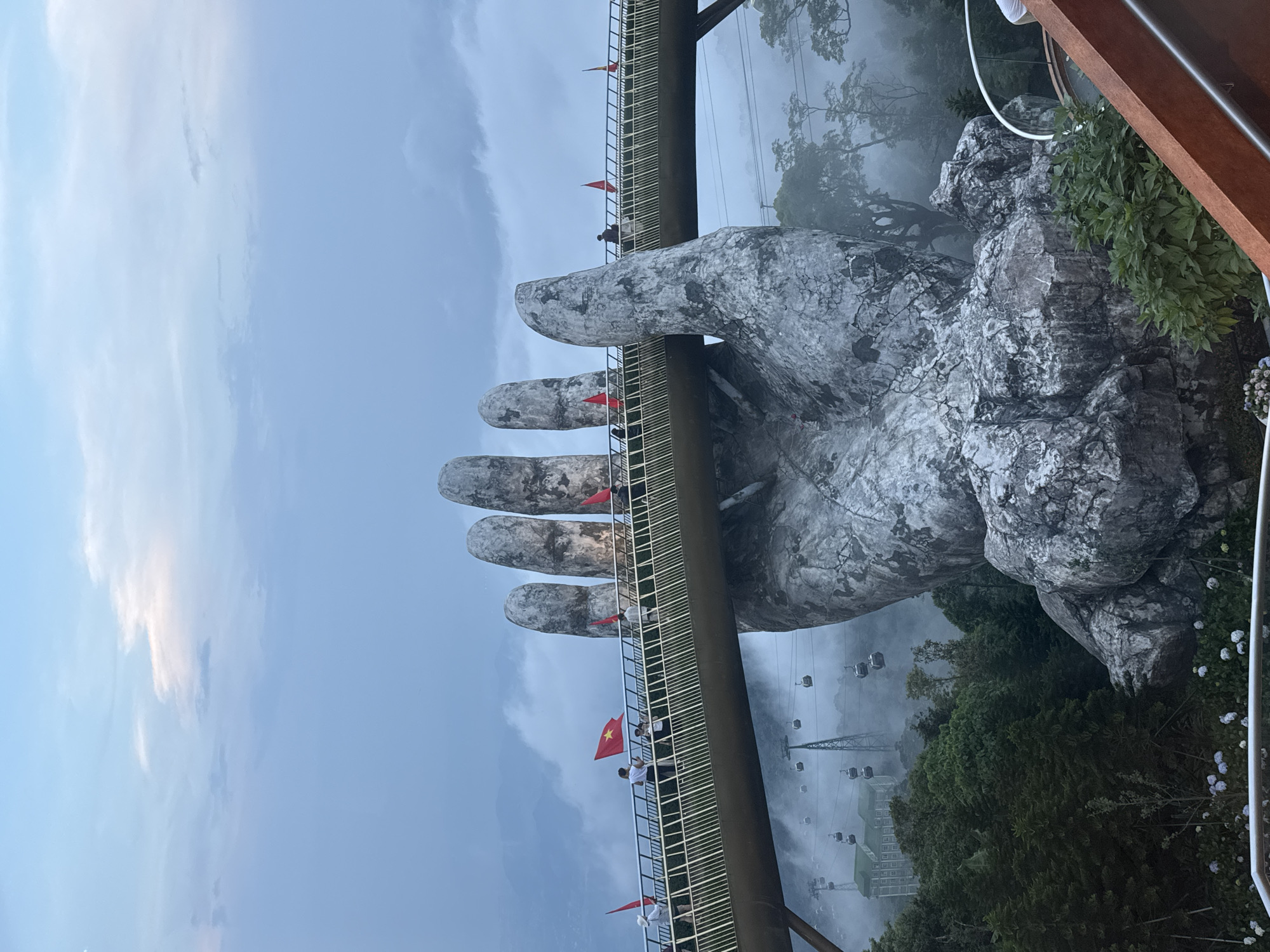 A panoramic shot captures the iconic Golden Bridge (Cầu Vàng) in Ba Na Hills, Da Nang, Vietnam, set against a misty, overcast sky. The bridge, with its distinctive golden-yellow hue and metal railings, appears to be cradled by two colossal, weathered stone hands that emerge dramatically from the landscape.
Several people, seemingly tourists, are visible strolling along the pedestrian bridge, some stopping to observe the panoramic views. Small, red Vietnamese flags with a yellow star are attached to the bridge's railing at regular intervals, adding a touch of national identity to the structure.
In the background, a verdant mountain valley is enveloped in a soft mist, creating an ethereal atmosphere. Numerous gondola cabins and their supporting pylons, part of a cable car system, are visible traversing the valley, disappearing into and emerging from the fog. Lush green trees and rugged rock formations are also present, especially around the base of the giant hands and along the mountain slopes. The lighting suggests daytime, possibly early morning or late afternoon, given the diffuse light and pervasive mist. The scene exudes a sense of wonder and grandeur, highlighting the unique architectural marvel within its natural, cloud-kissed environment. No visible text is present in the image.