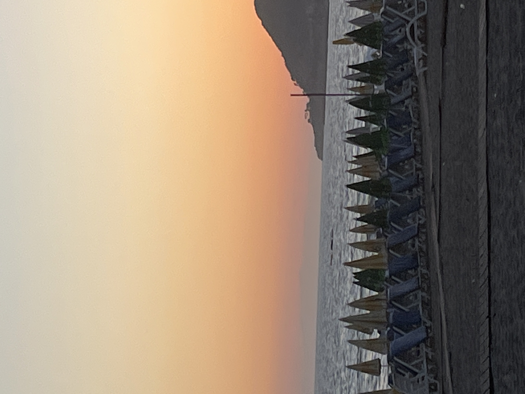 The image shows a row of beach umbrellas and lounge chairs on the shoreline of Alanya, Turkey. The sky is a gradient of orange and yellow, suggesting sunset or sunrise. A dark, mountainous landmass is visible in the background. The water reflects the colors of the sky. The beach umbrellas are various shades of yellow and green. A wooden pier or boardwalk runs along the right side of the image.