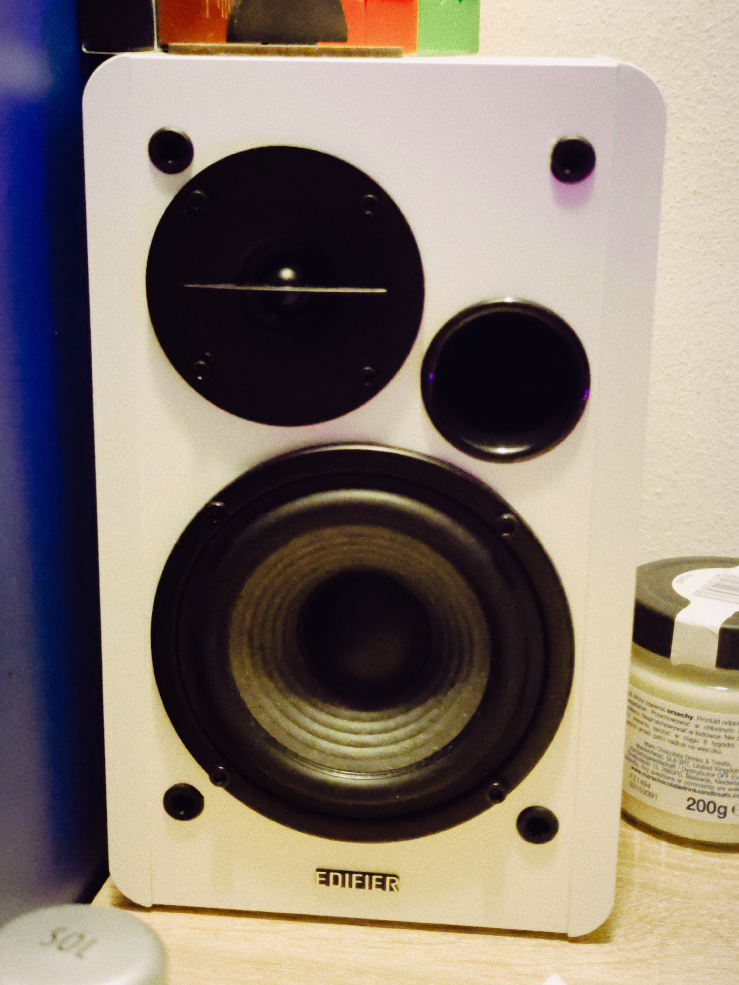 The image shows a white Edifier speaker with black speaker cones. The speaker has two smaller cones at the top and a larger one at the bottom. The Edifier logo is visible on the bottom of the speaker. To the left, there is a partial view of a blue object and the lid of a jar is at the bottom. To the right of the speaker is a 200g jar with visible writing. The exact location in Budva, Montenegro, is impossible to determine from the given image.
