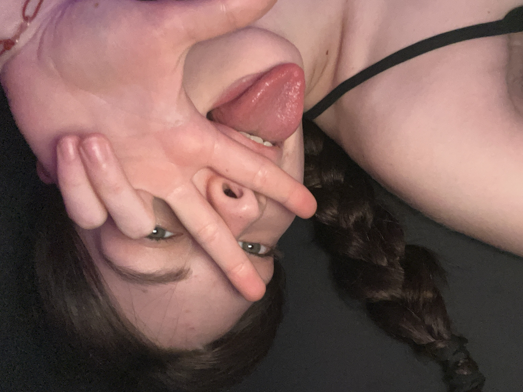 A close-up, upside-down, low-angle shot depicts a fair-skinned person with light eyes and brown hair, featuring a single braid. The person is sticking out their tongue while partially covering their face with a hand. The background appears to be a dark, solid color. A thin black strap, possibly from clothing, is visible on the upper right. No location is identifiable from the image.