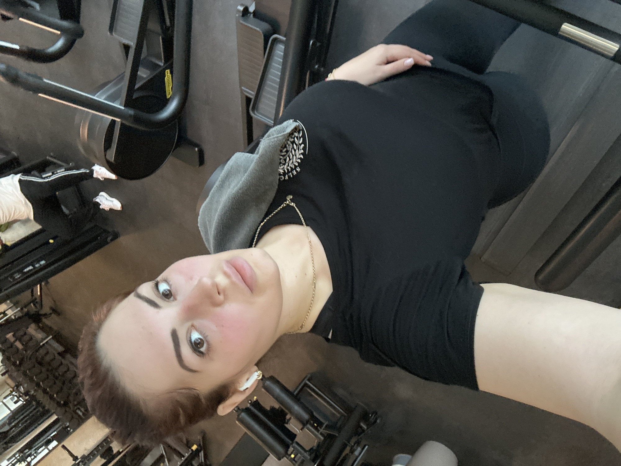 A woman is posing for a selfie in a gym. She is wearing a black t-shirt, black leggings, and an earbud in her right ear. A gray towel is draped over her shoulder. She is wearing a gold chain necklace and a bracelet on her left wrist. Gym equipment, including treadmills and weight machines, is visible in the background. The gym's specific location in Kazanlak, Bulgaria is not discernible from the image.