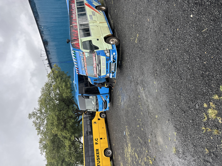 Three buses are parked in a gravel lot in front of a blue building. The buses are all facing the same direction, with the closest bus being a white and blue Isuzu minibus with the words "S.M.P.E.C. " and "Suzuki Champion" on it. Behind the minibus is a blue and white bus with a faded "TUSKER F.C." on its side. The furthest bus is a blue and white bus. A person in a black shirt stands next to the closest bus. There is a large green tree in the foreground of the image. The image is taken in the industrial area of Machakos, Kenya, at latitude -1.52 and longitude 37.264.