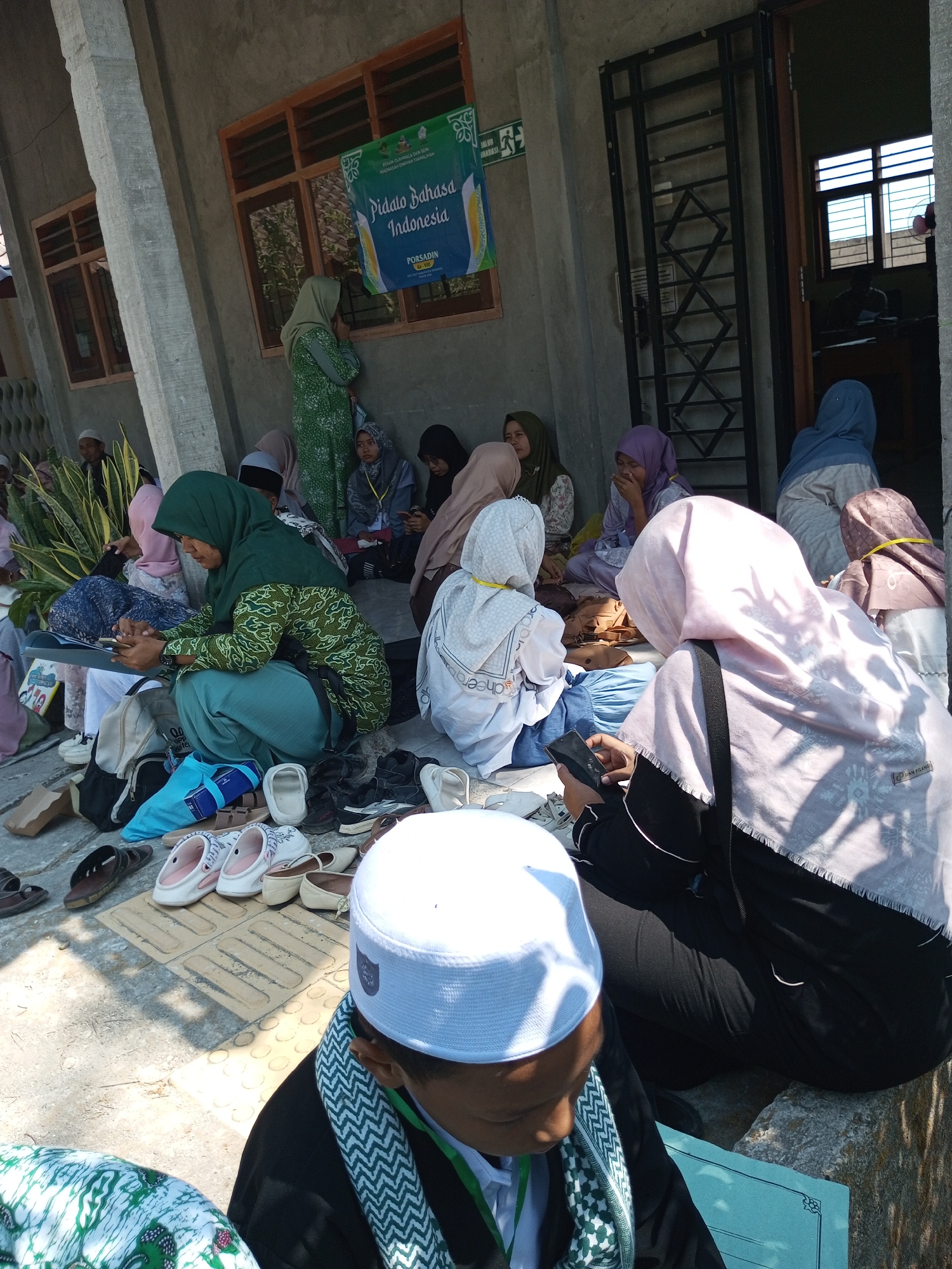 The image depicts a gathering of people, predominantly women wearing hijabs, seated on the ground outside a building. The setting appears to be in Dresi Kulon, Indonesia, as indicated by the presence of a banner that reads "PORSADIN Pidato Bahasa Indonesia Dresi Kulon, Tahun 2020." Some individuals are engaged with their mobile phones, while others appear to be conversing or resting. Various footwear, including sandals and shoes, are scattered on the ground around the seated individuals. The building has concrete pillars, a metal-barred door, and windows with wooden frames. The overall atmosphere suggests a communal event or religious gathering.