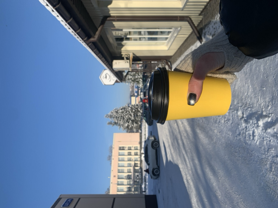 A first-person perspective photograph captures a right hand holding a bright yellow disposable cup with a black lid. The hand wears a light-colored ribbed fingerless glove and a dark jacket sleeve, with nails painted in a dark, glittery polish. The scene depicts a sunny winter day in Bohuslav, Ukraine.
The foreground consists of snow-covered ground with visible footprints and long shadows cast by unseen objects. To the immediate right, the exterior of a light-colored building with vertical siding, a brown drainpipe, and a window with white frames is visible. An outdoor air conditioning unit is mounted on this building. Above the building's roofline, a white circular sign featuring a black coffee cup icon is present.
In the mid-ground, a snow-covered parking area contains several vehicles, including a white SUV prominent on the left and other darker vehicles further back. Two snow-laden evergreen trees are situated between the vehicles and another distant building. Further back on the left, part of a light-colored building with a blue sign bearing Cyrillic text (appears to read "ШОН") is visible. Beyond this, a larger, multi-story pinkish-beige building with numerous windows stands. The sky is clear and bright blue.