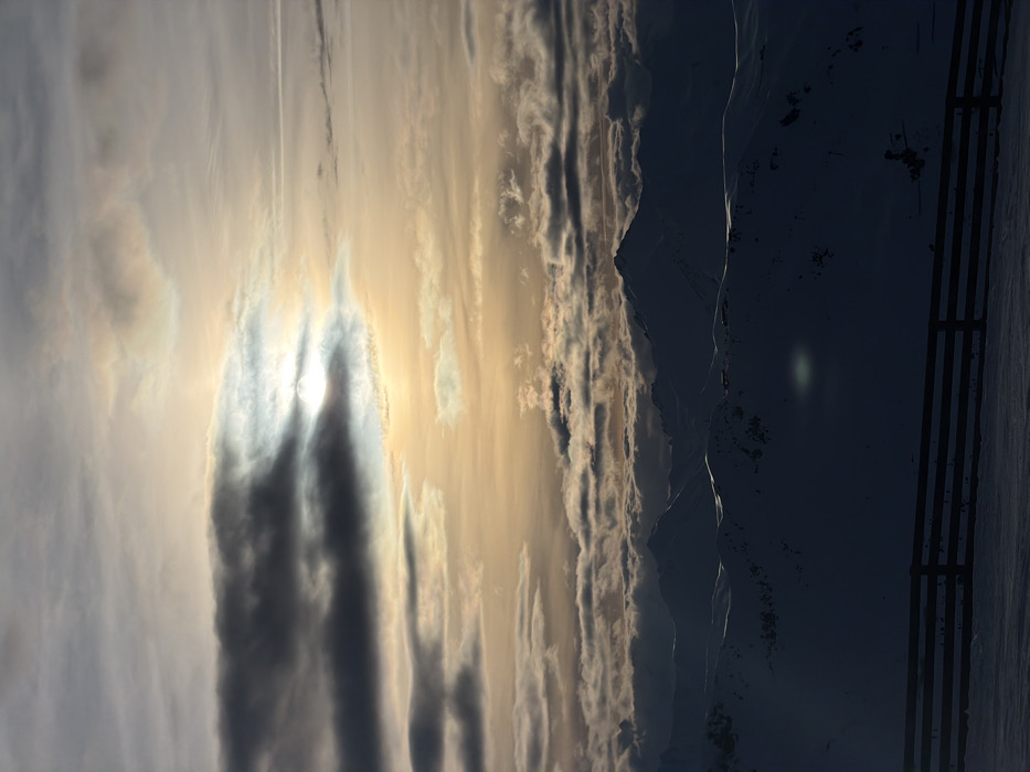 Mountains under a sky streaked with clouds. Sunlight filters through a break in the clouds creating an ethereal effect. A dark wooden fence runs vertically along the right edge, juxtaposed against the natural scenery. The overall scene presents a serene yet powerful vista, dominated by the mountain range and the expansive, cloud-filled sky.