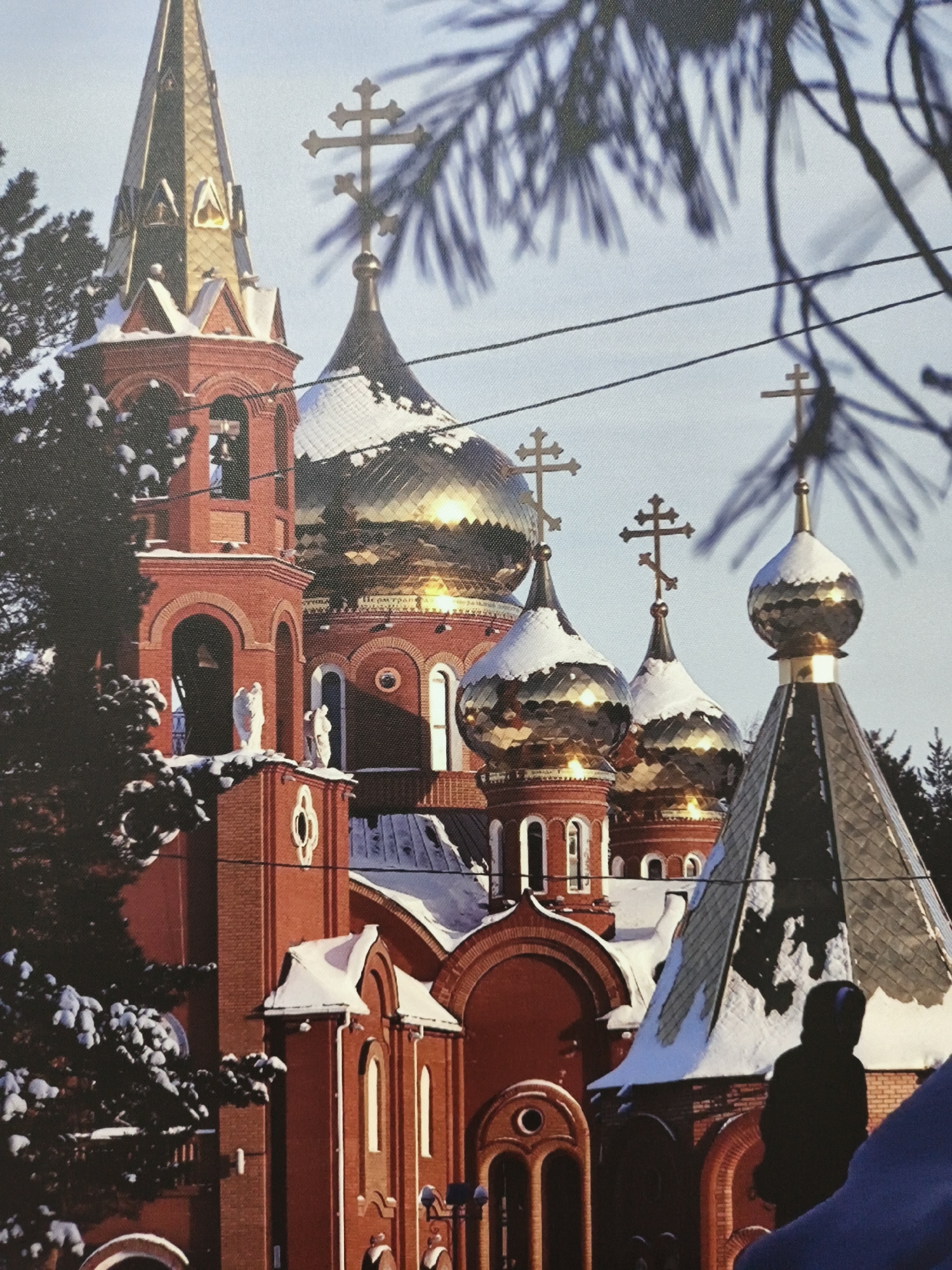 A picturesque winter scene captures a snow-dusted Eastern Orthodox church complex, dominated by rich red brick architecture and numerous glistening golden domes. The prominent central dome reflects bright sunlight, and several smaller, ornate onion domes are also covered with snow at their bases. Each dome and spire is topped with an Orthodox cross.
On the left, a tall, golden pointed spire rises from a red brick bell tower, where bells are visible within its arched opening. Below the largest dome, two white statues, possibly angels, stand on a ledge. Arched windows and decorative brickwork are visible throughout the structure. Dark, snow-laden evergreen tree branches blur in the foreground and frame the top and right side of the image, adding depth. Thin power lines stretch across the clear, bright sky. On the lower right, a dark silhouette of a person, partially obscured, is visible. Faint, highly reflective text, possibly Cyrillic, is discernible on the largest golden dome, but it is not clearly legible. The scene suggests a cold, clear winter day, highlighting the architectural beauty of the church.