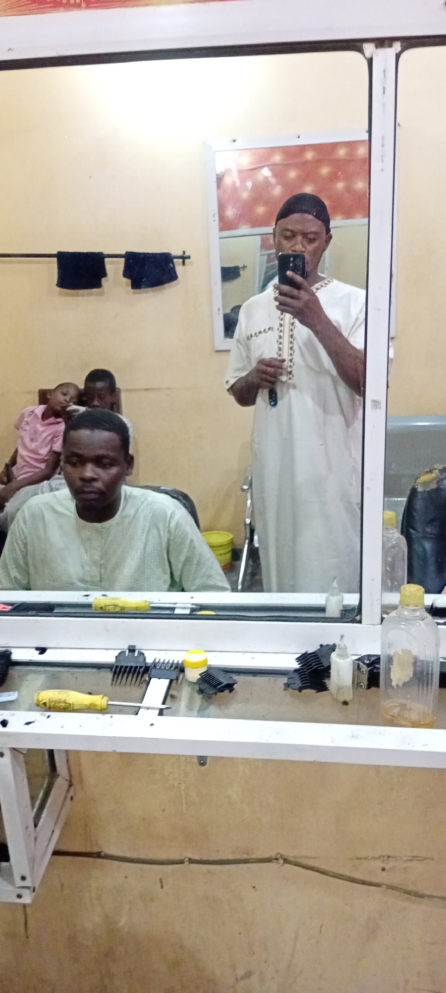 The image shows the interior of what appears to be a barber shop in an unknown location.
A man in a light green garment is sitting in the barber's chair. Behind him, two children are seated. Another man in a white robe and a black cap stands in the reflection of a large mirror, holding a smartphone, seemingly taking a selfie. On the surface in front of the mirror are various barbering tools including combs, clippers, and bottles. The room has pale yellow walls, and a clothes rail with a couple of dark blue towels hangs in the background.