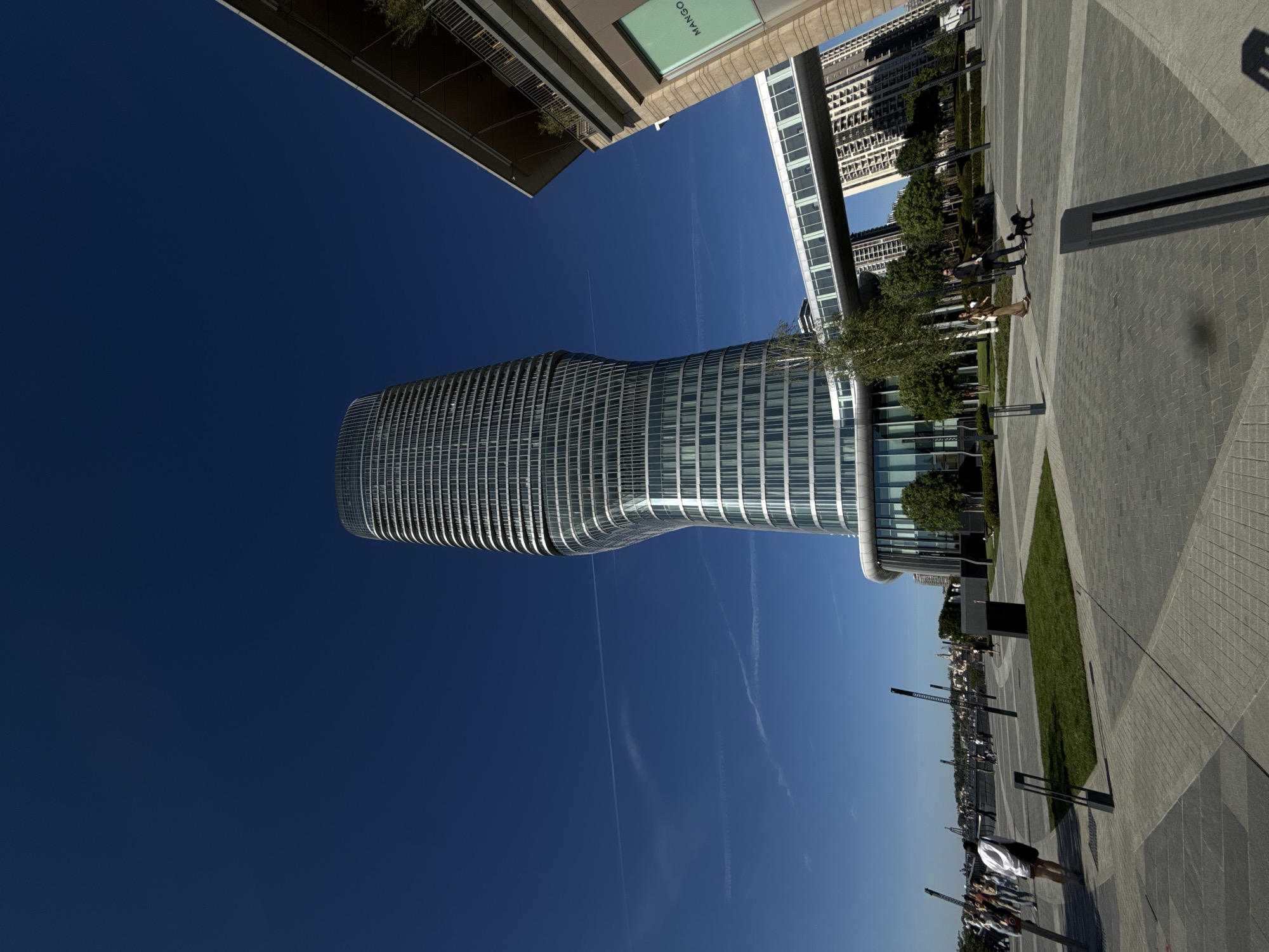 The image presents a cityscape with a modern architectural emphasis, taken from a low-angle perspective. Dominating the frame is a curved, glass-paneled office building, its horizontal striations suggestive of a contemporary design. The building is set against a deep blue sky, contrasting with the building's reflective surface. Adjacent to the office building are other structures, including what appears to be a shopping complex with signage for "Mango."
The ground-level presents a public space with geometric paving and patches of green lawn. There are individuals walking, some accompanied by pets, suggesting an active pedestrian environment. Street furniture, like lighting fixtures, is also visible. The background features a distant view of what could be the older sections of Belgrade, indicating the juxtaposition of modern and historic elements in the city. The image suggests that this could be the Belgrade Waterfront development area, a modern project in the city.
