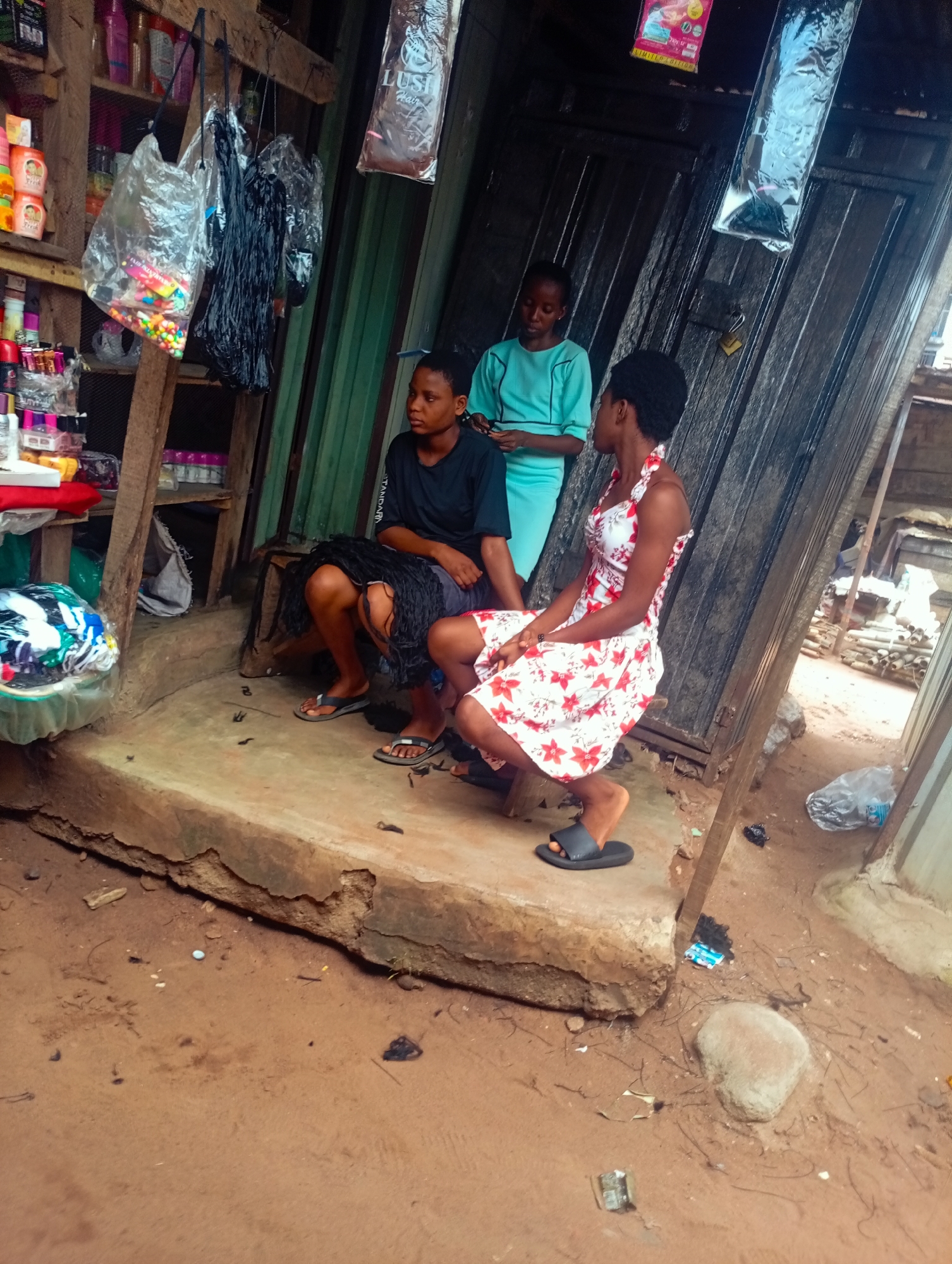 Three young women are sitting on a raised concrete ledge in front of what appears to be a small market stall. The stall is made of rough wooden planks and is open to the elements. Shelves are visible inside, stocked with various items including bottles and packaged goods. Hanging from the ceiling are plastic bags containing colorful beads and what look like hair extensions.
The woman on the left is wearing a black t-shirt and a dark skirt or wrap, with long black braided hair. She is looking to her left. The woman in the middle is wearing a light blue, short-sleeved tunic-style top. She is looking down at something in her hands. The woman on the right is wearing a white and red floral print dress. She is looking to her left. All three women are wearing flip-flops.
The ground in front of the stall is dirt and appears somewhat uneven, with scattered debris. There are also some objects on the ground, including a plastic bag and a rock. The scene suggests a warm, possibly sunny day, given the lighting and the attire of the individuals. The overall impression is one of a casual gathering at an outdoor marketplace. There are no visible signs with legible text.