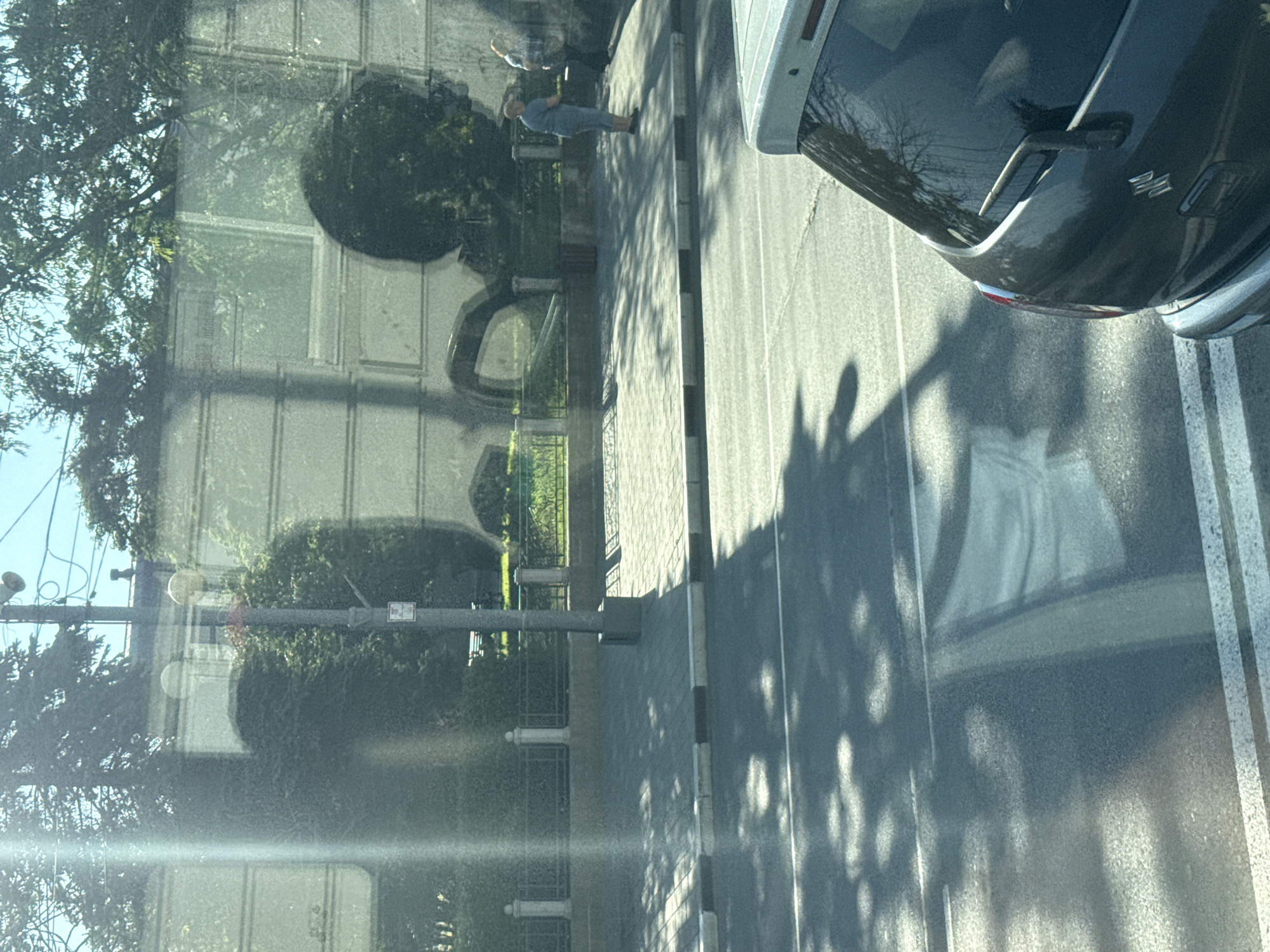 The image, taken from inside a vehicle, shows a street scene in Yuzhno-Sakhalinsk, Russia. In the foreground, the rear of a dark-colored hatchback car is visible, with its rear window and wiper present. The road surface is asphalt, marked with white lines, and dappled with shadows from overhead trees. To the left of the road is a paved sidewalk with a black and white striped curb. Beyond the sidewalk is a low fence with white decorative pillars, behind which are manicured bushes and trees. In the mid-ground, a lamppost with a circular light fixture stands on the sidewalk. A light-colored building with horizontal and vertical lines is visible behind the foliage. Two individuals are standing on the sidewalk near the fence, one wearing a blue shirt and light-colored pants, and the other in a plaid shirt. Reflections are visible on the building and the car's rear window, suggesting the photograph was taken through glass. The overall lighting indicates it is daytime, with bright sunlight casting distinct shadows.
