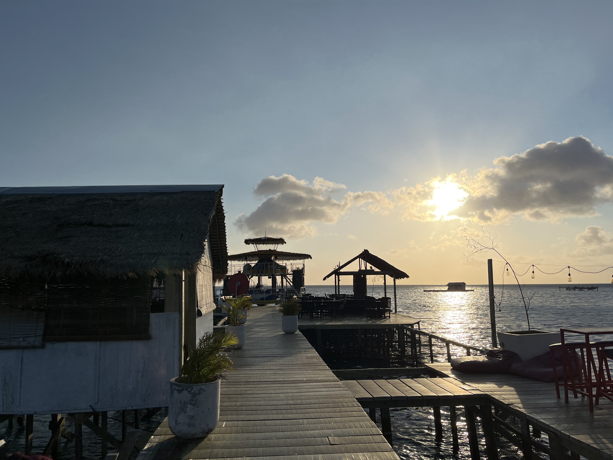 A wooden boardwalk extends into a calm ocean at sunset, leading to several stilted structures with thatched roofs. The boardwalk has planters with greenery and some seating areas. In the distance, a small floating structure and a distant island are visible. The sky is a clear blue with scattered clouds, and the sun is setting, casting a warm glow on the water. The scene appears to be a resort or restaurant in Karimunjawa, Indonesia, known for its beautiful beaches and marine life.