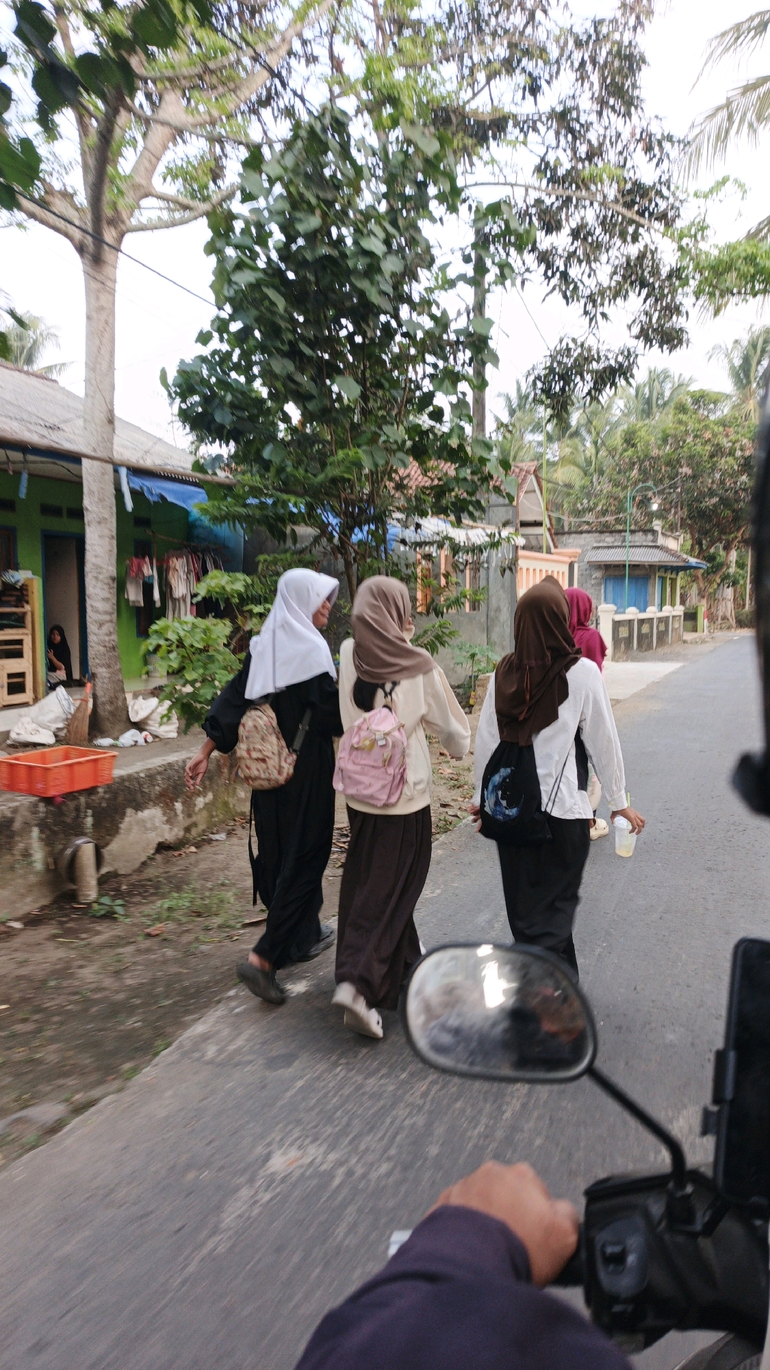 A low-angle, wide shot captures three young women walking on a paved road in Cibenda, Indonesia. The woman on the left wears a white hijab and a black abaya, carrying a patterned backpack. The middle woman has on a light brown hijab, a cream-colored long-sleeved top, and a dark brown skirt, with a pink backpack. The woman on the right, further down the road, wears a brown hijab and a white long-sleeved top, carrying a black backpack and a clear plastic cup. The road is flanked by modest houses with colorful facades, one with laundry hanging outside. Lush green trees with some bare branches are visible behind the houses. The perspective suggests the photo is taken from a motorcycle, as a hand and part of a motorcycle mirror are visible in the foreground. The lighting suggests it is daytime.